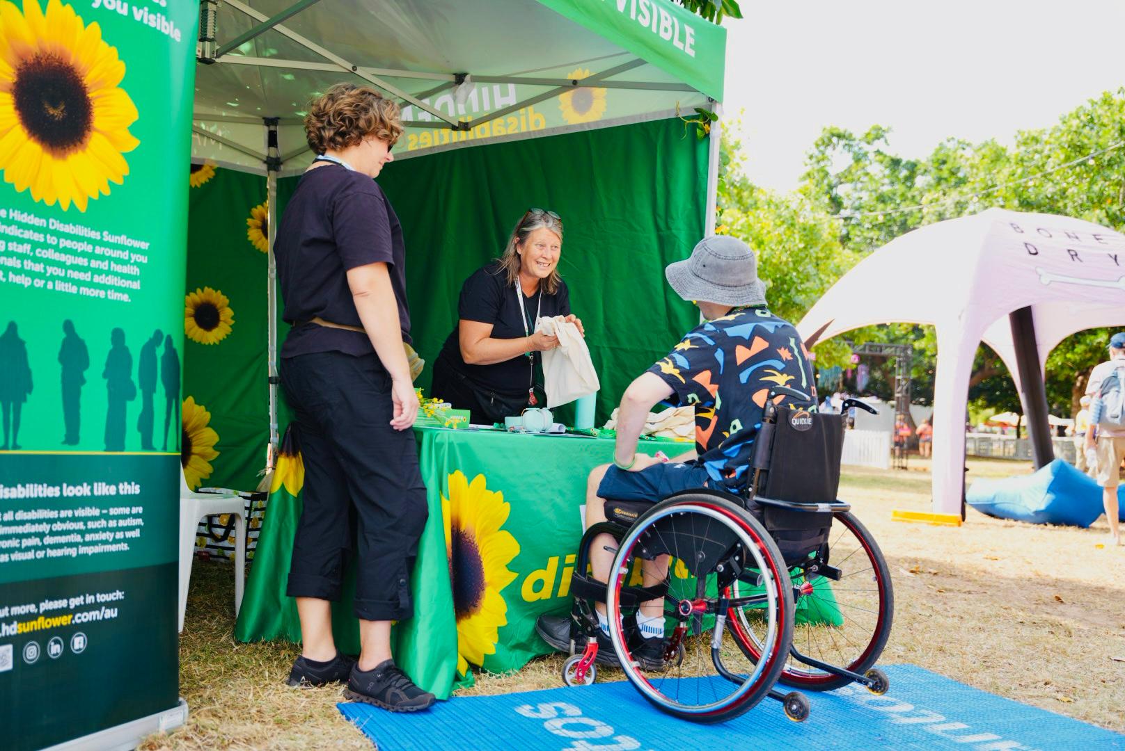 A person in a wheelchair interacts with two people at a sunflower-themed booth in an outdoor setting.