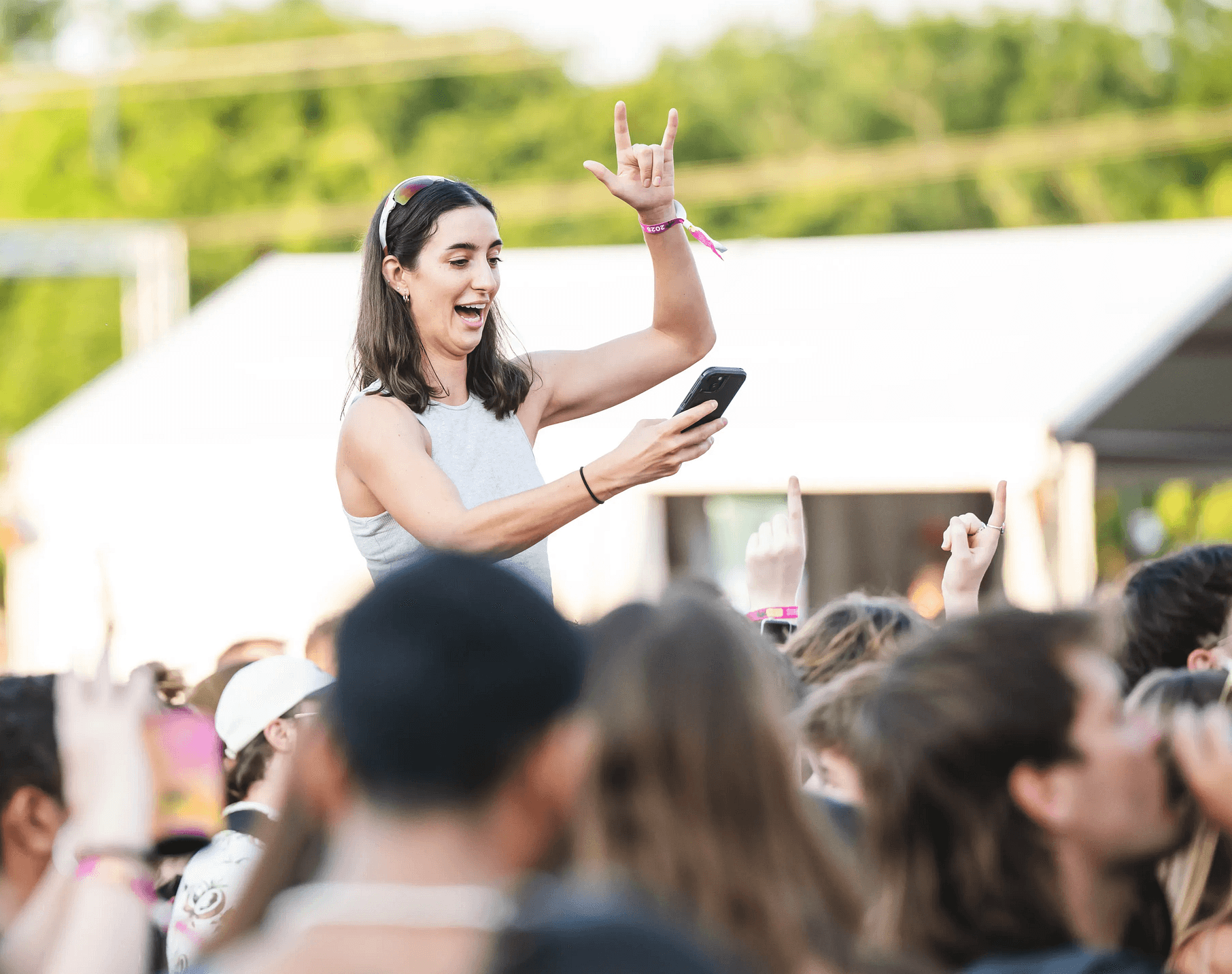 A woman on someone's shoulders at an outdoor concert, smiling and holding a phone, with people around her and trees in the background.