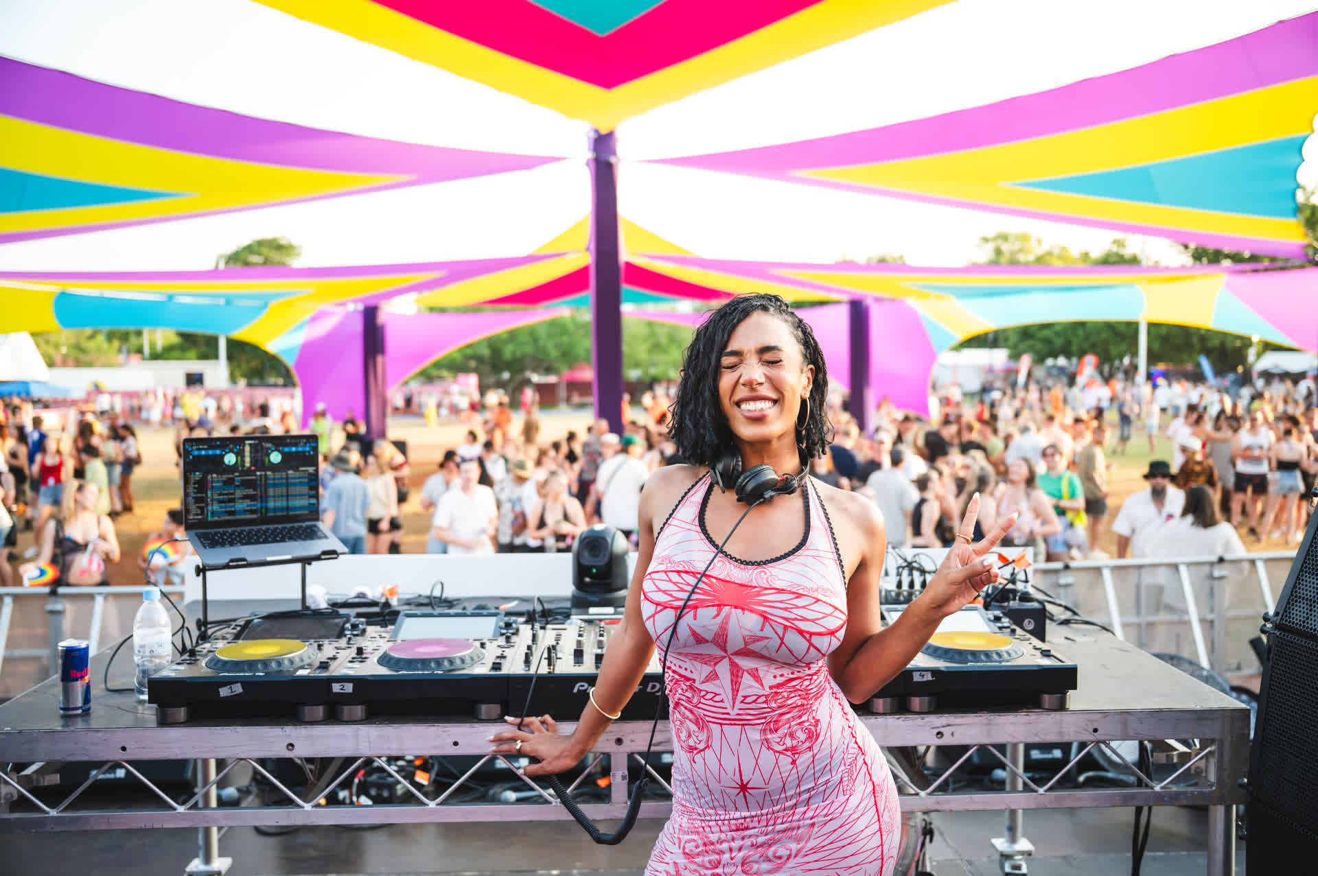 DJ smiling at an outdoor festival, standing in front of a colorful stage with vibrant canopy and a crowd in the background.