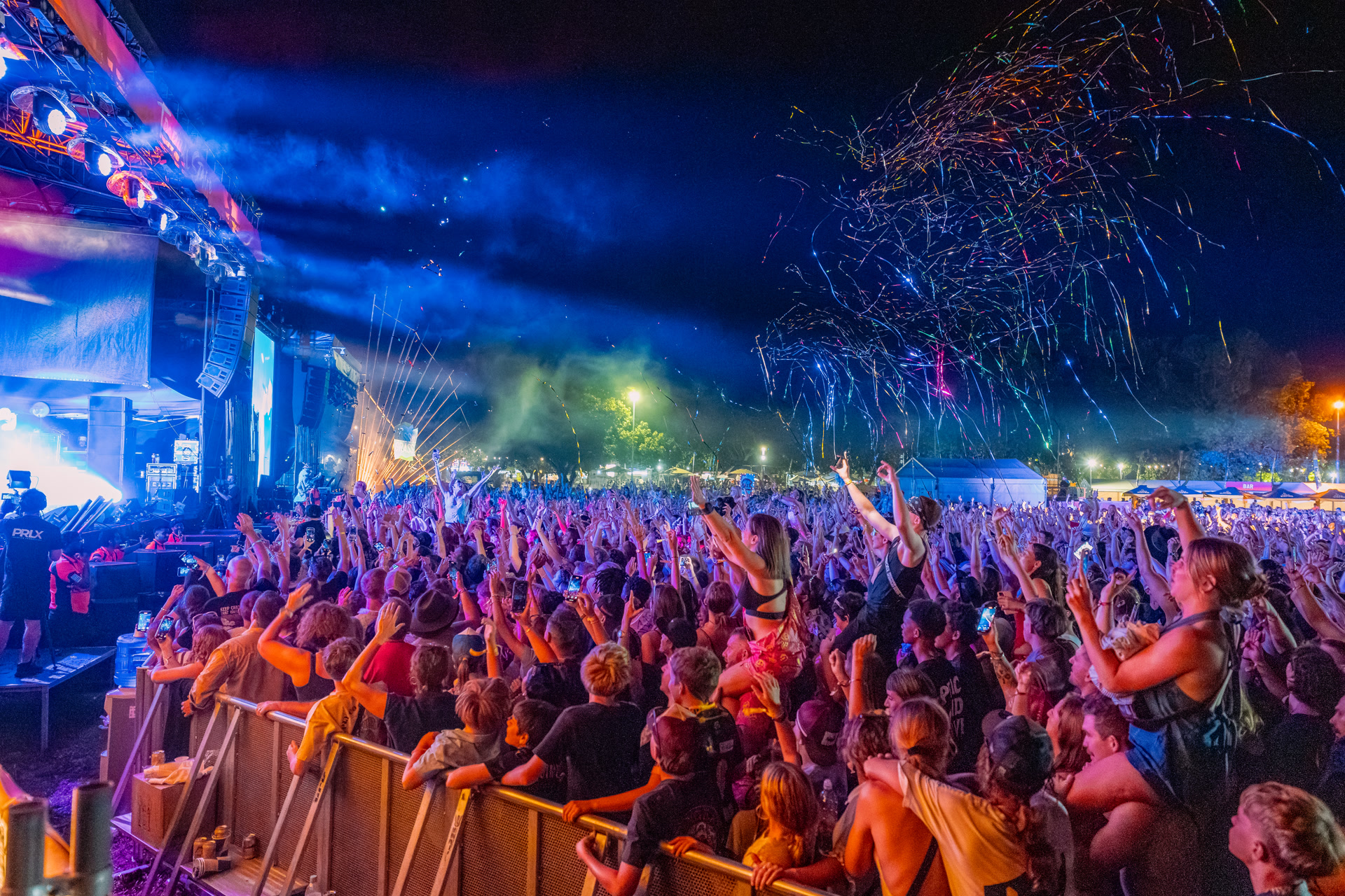 A lively outdoor concert at night with a cheering crowd, confetti in the air, and colorful stage lights illuminating the scene.