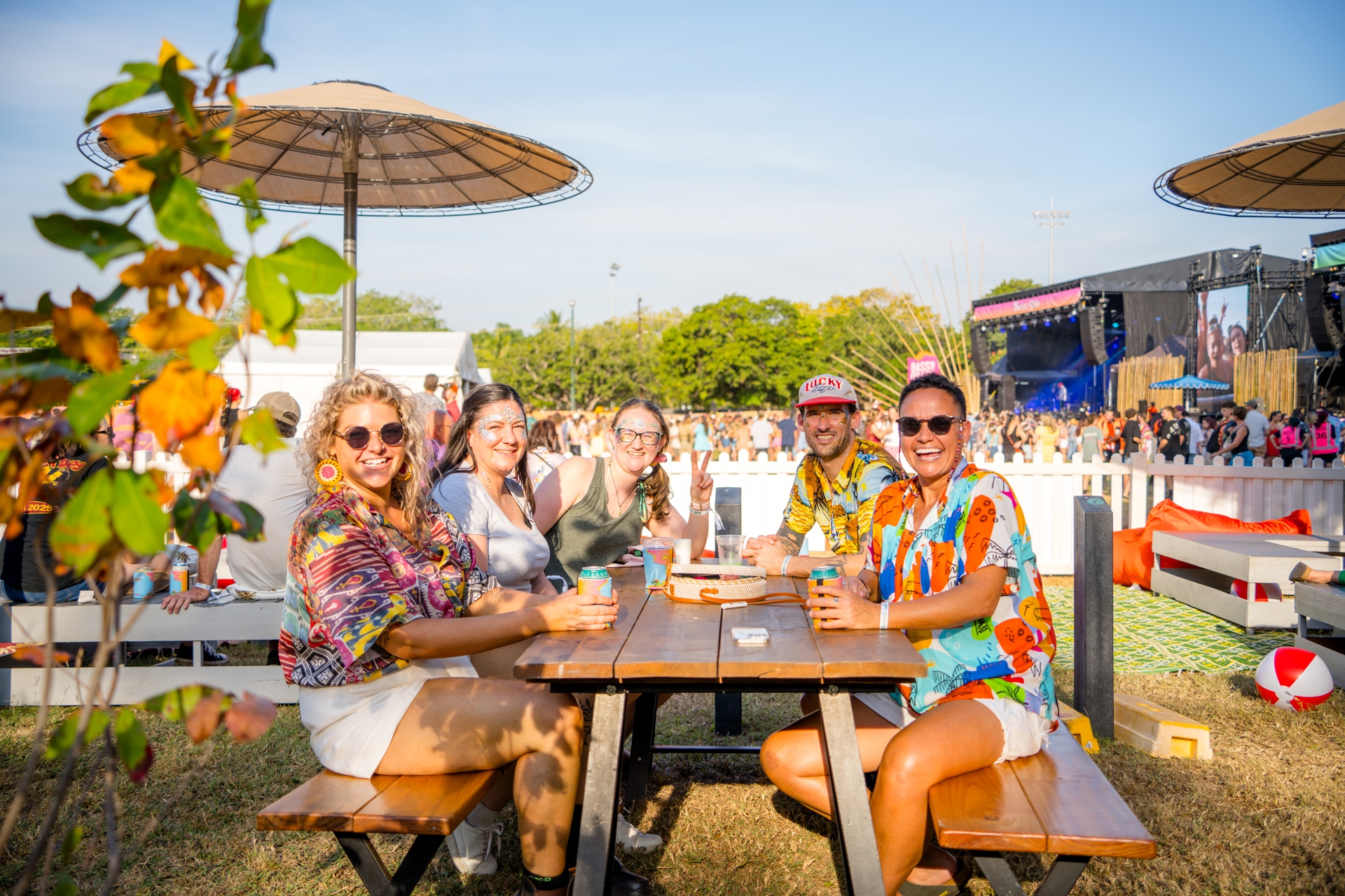 A group of people sitting at a picnic table outdoors, wearing colorful clothes, smiling, with a stage and crowd in the background on a sunny day.