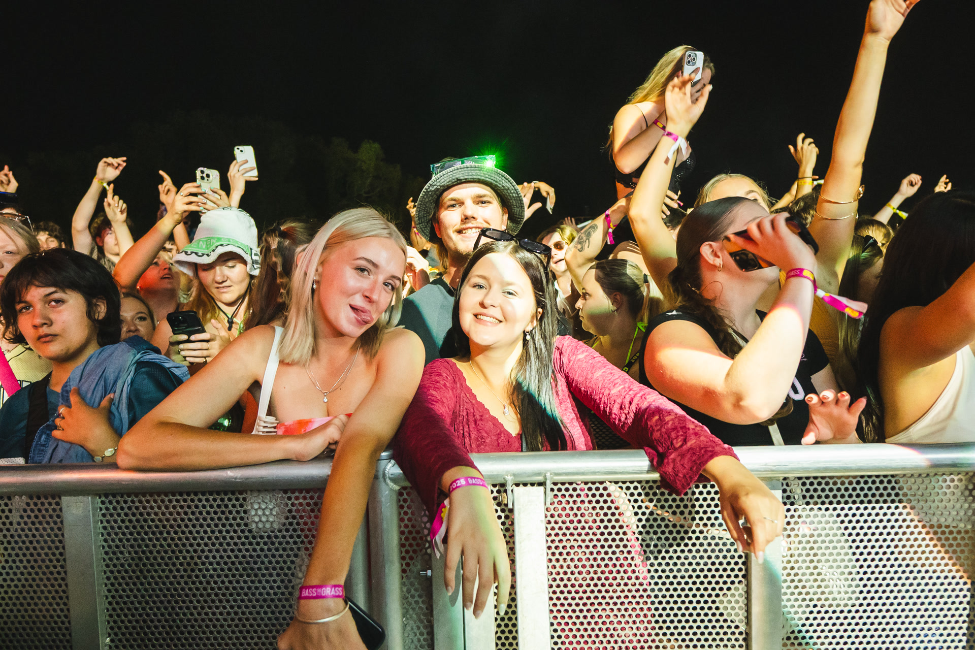 A lively crowd at a concert, with people smiling, cheering, and taking photos. Bright stage lights illuminate the scene.