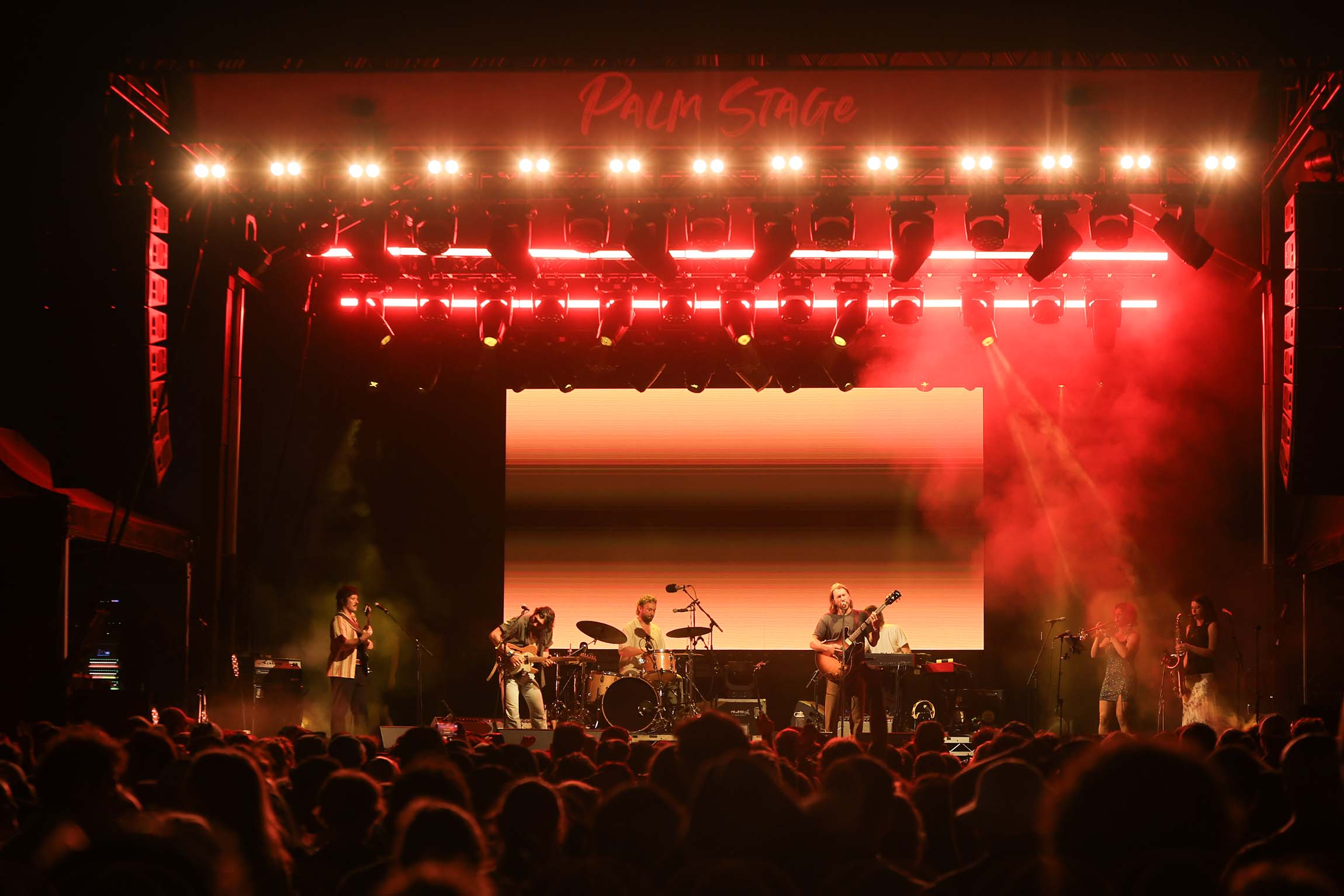 Band playing on "Palm Stage" under intense red lights, with a large crowd. Features guitar, drums, trumpet, and saxophone.