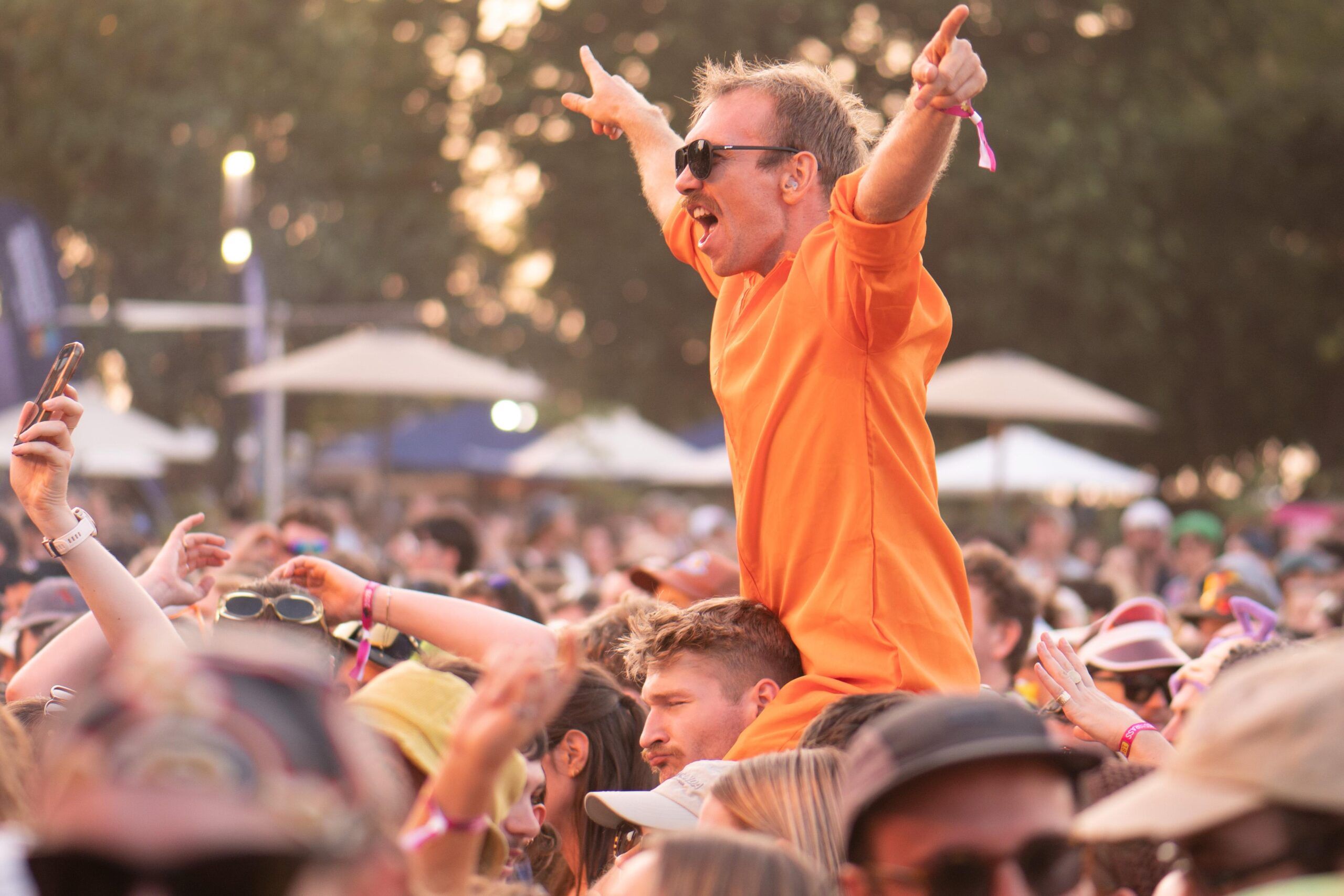 Man in orange shirt on someone's shoulders, cheering at an outdoor festival, surrounded by a lively crowd.