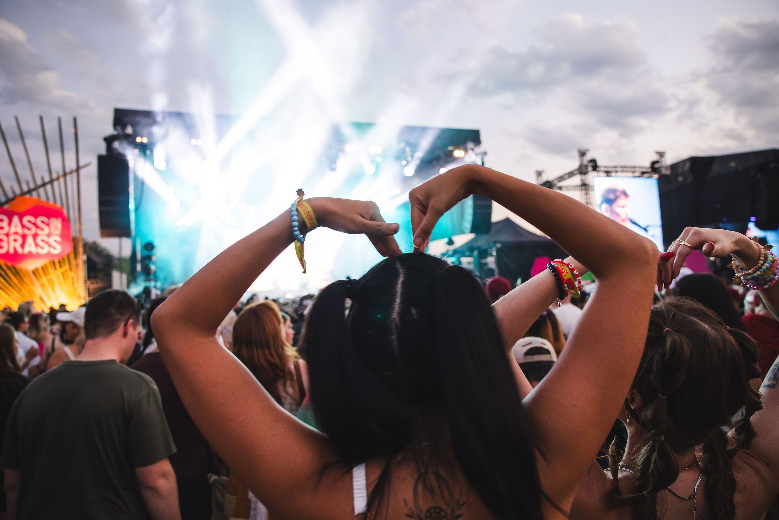 Concertgoer with braided hair forms a heart with hands, surrounded by a lively crowd, colorful wristbands, and a brightly lit stage.
