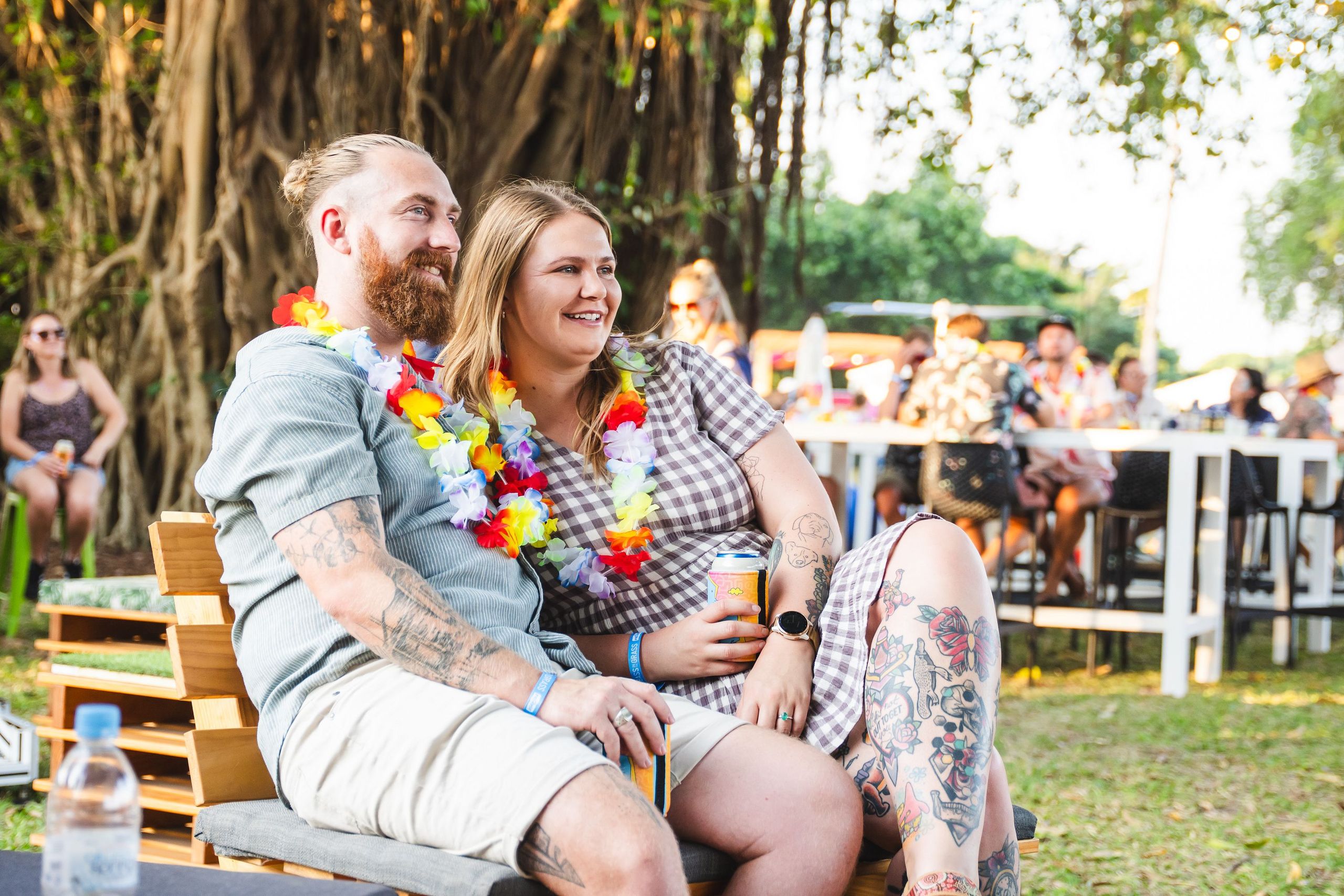 A smiling couple with leis sits on a wooden bench at an outdoor event, surrounded by people, trees, and tables.