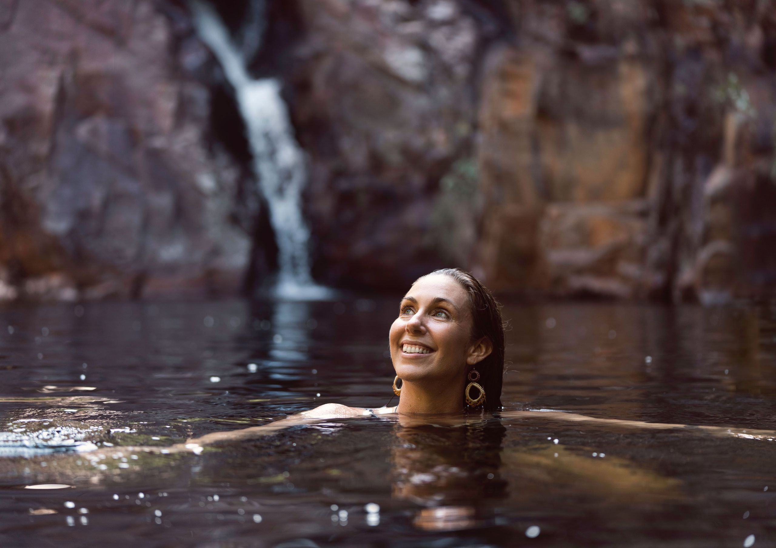 Woman smiling while swimming in a serene natural pool with a waterfall and rocky backdrop.