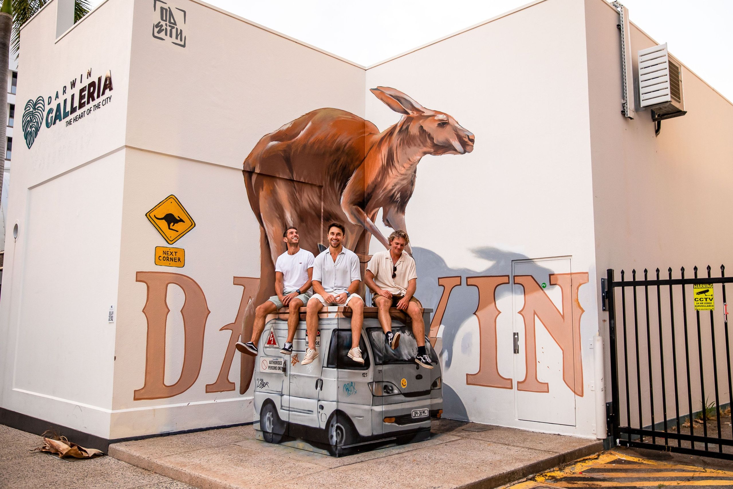 Three people sitting on a small vehicle in front of a building with a large kangaroo mural and "Darwin" text.
