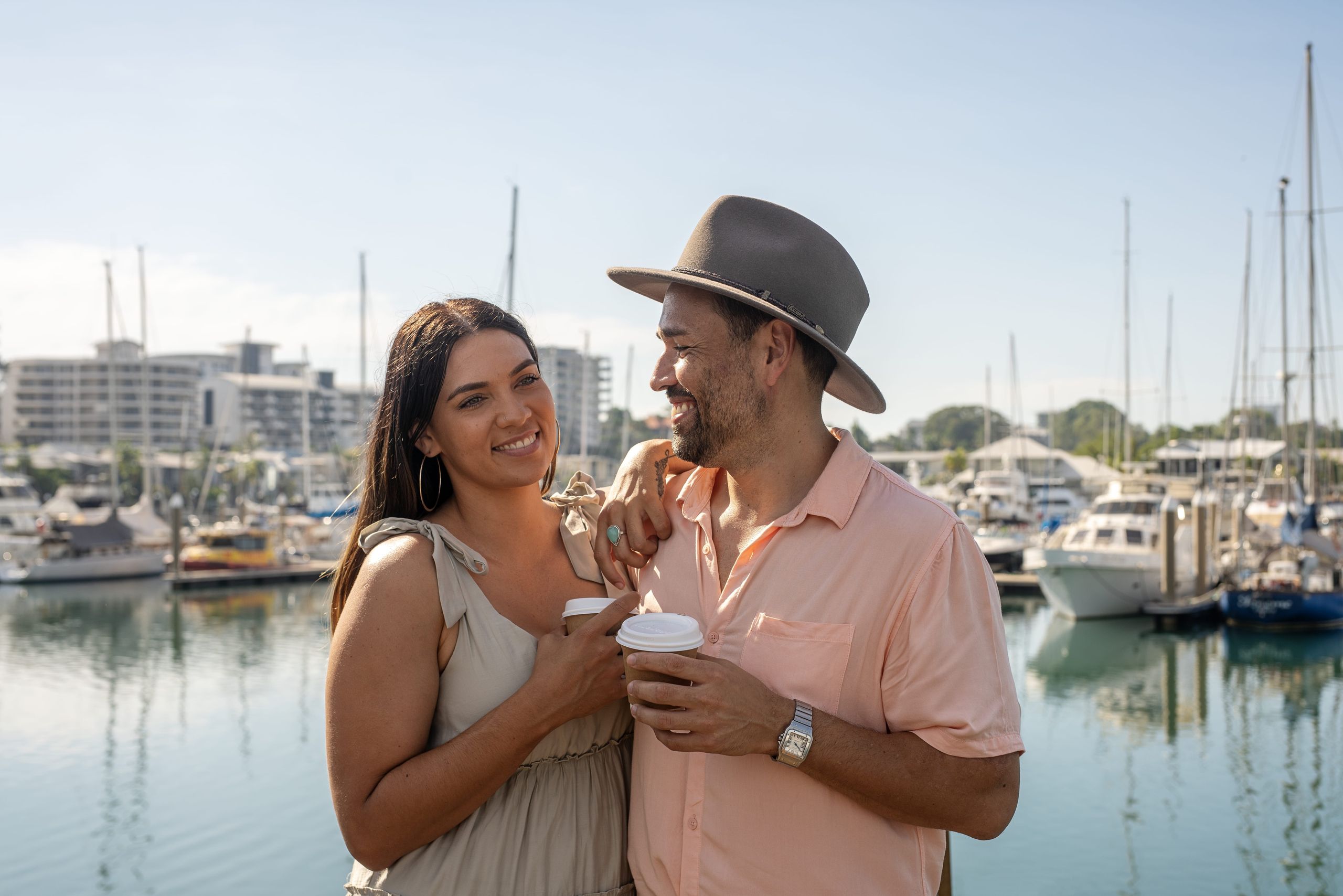 A smiling couple stands by a marina, holding coffee cups. The woman wears a beige dress, and the man wears a hat and peach shirt. Sailboats are in the background.
