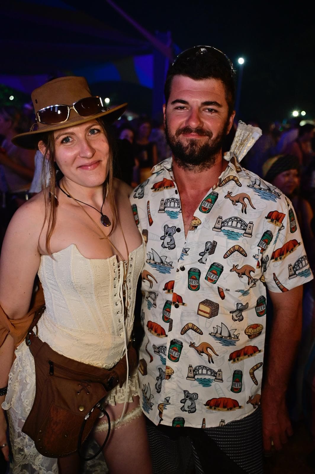 A smiling woman in a white corset and brown hat stands next to a bearded man in a patterned shirt at an outdoor event.