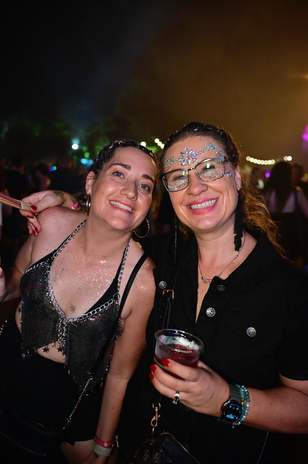Two women smiling at a nighttime event, wearing glittery outfits and face decorations, one holding a drink.