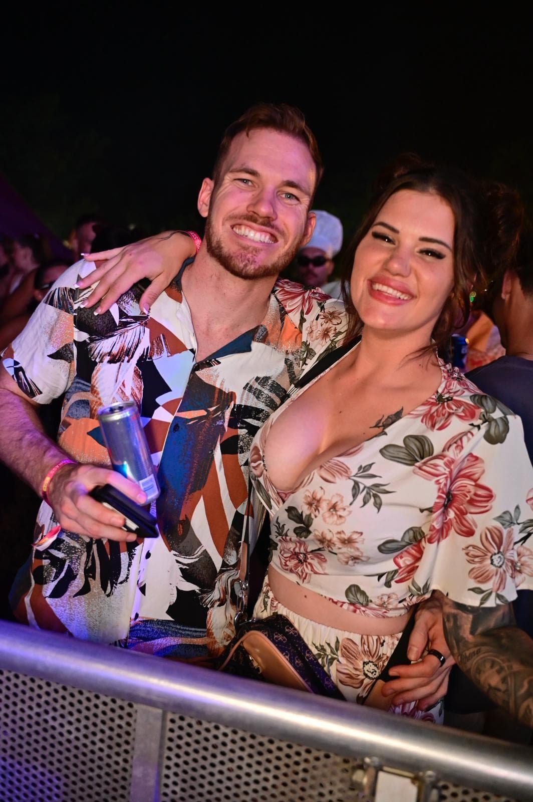 Smiling couple in floral outfits at an outdoor event, with the man holding a drink and the woman resting her arm on his shoulder.