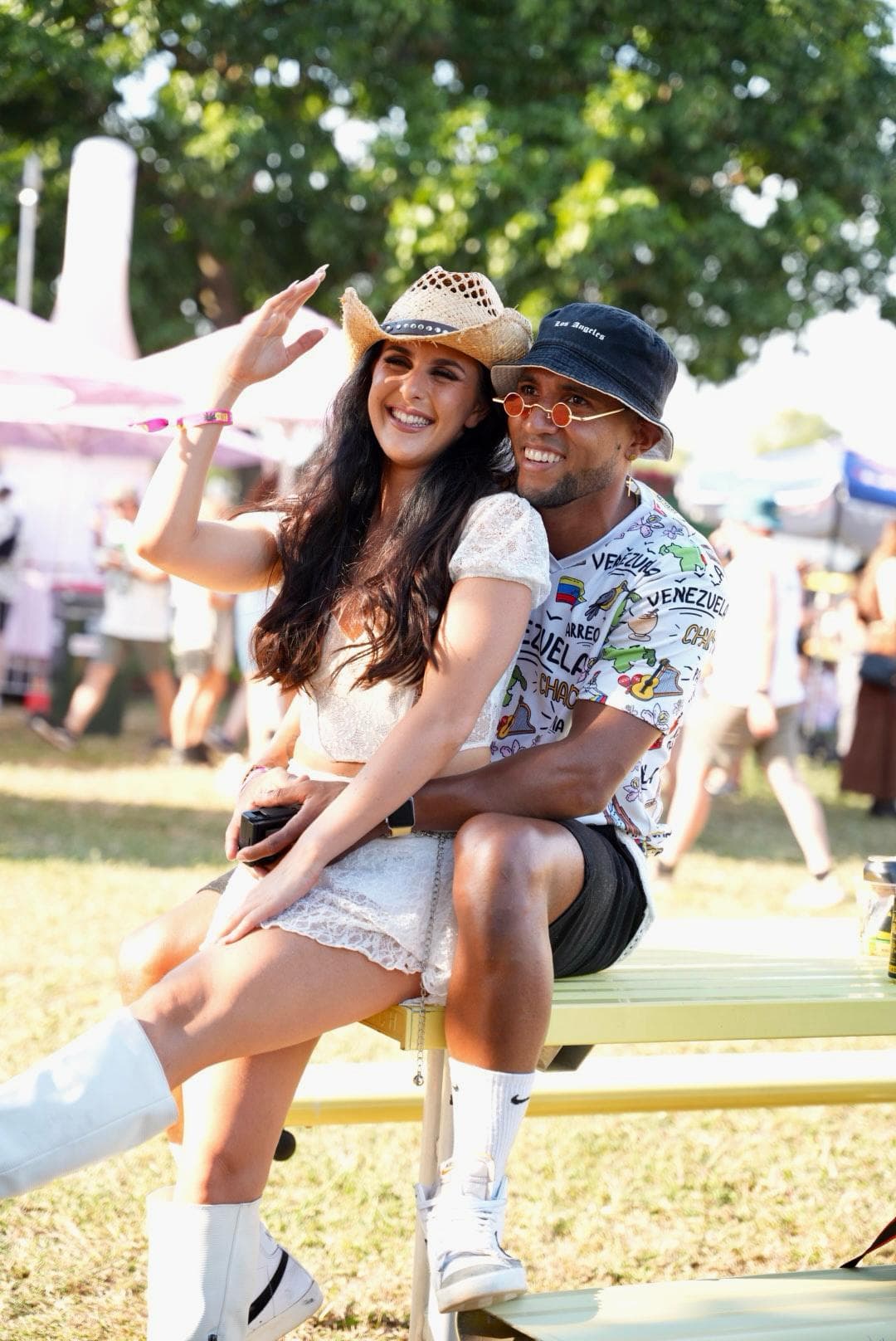 A smiling couple sits on a picnic table at an outdoor event. The woman wears a cowboy hat and boots, and the man wears a printed shirt and bucket hat.