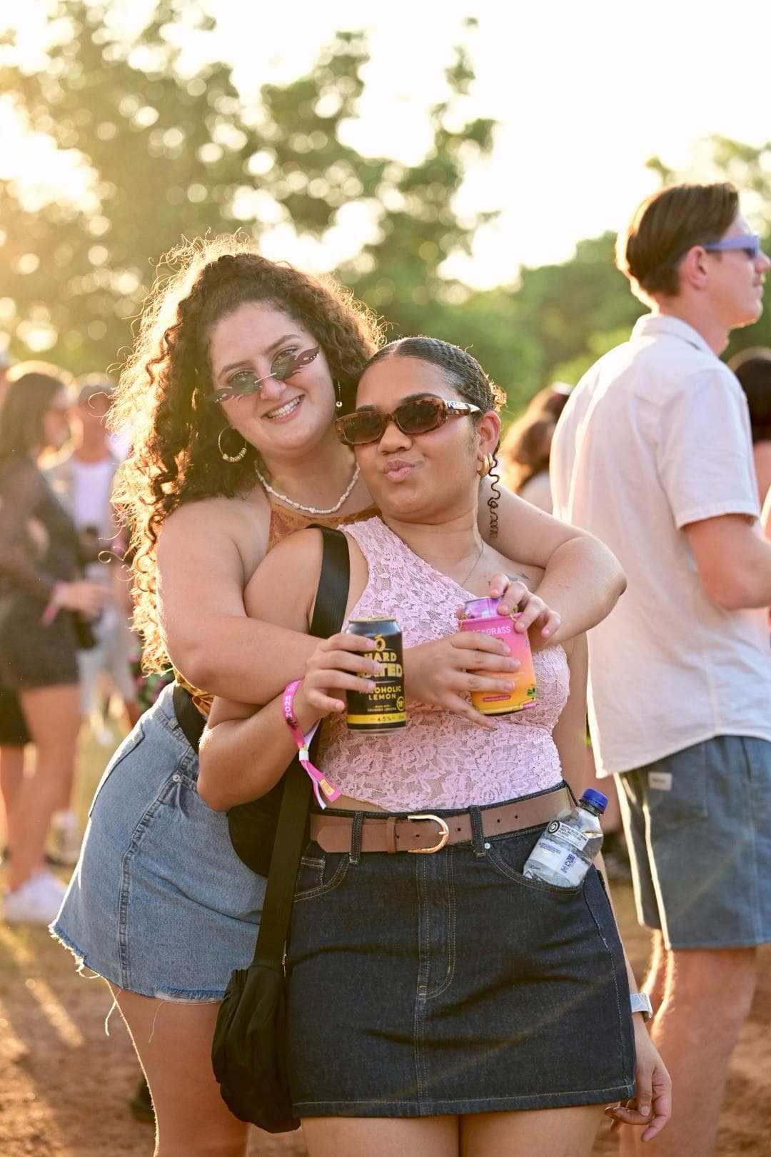 Two people smiling, embracing, and holding drinks at an outdoor event. They are wearing sunglasses and casual summer clothing.