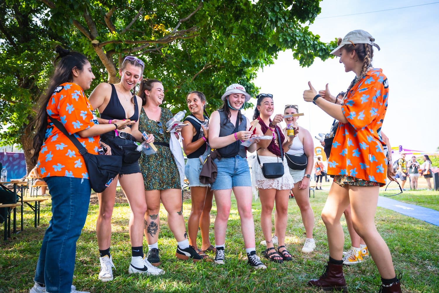 A group of smiling people stand together outdoors, some holding drinks. One person gestures with thumbs up. They are under a tree on grass.