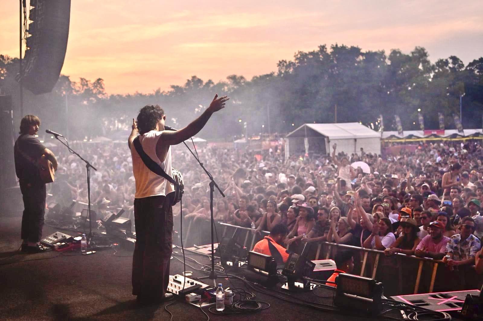 Musicians perform on stage at an outdoor concert, with a large crowd and colorful sunset sky in the background.