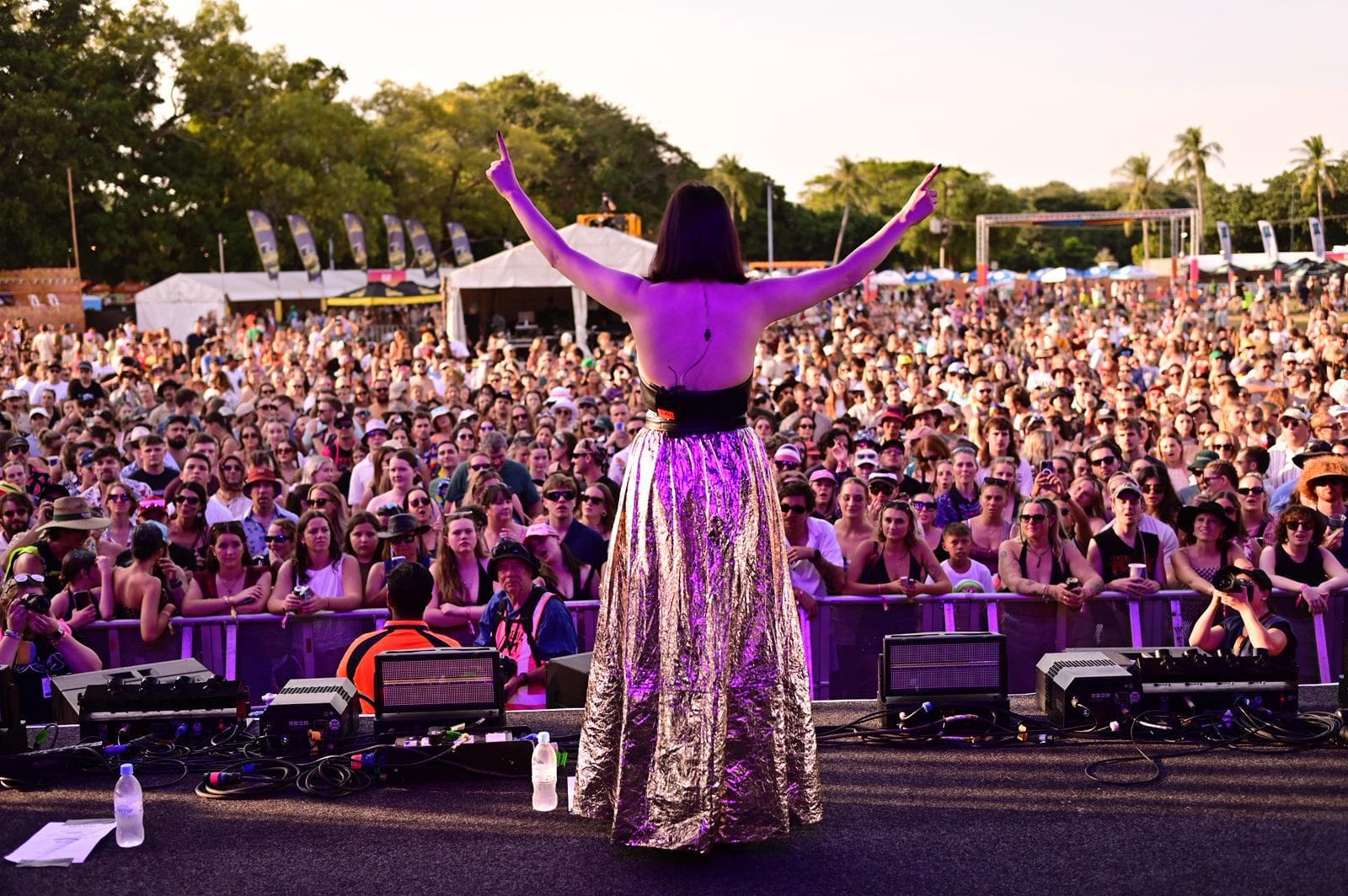 A performer in a shiny skirt faces a large festival crowd from the stage, with trees and tents visible in the background.