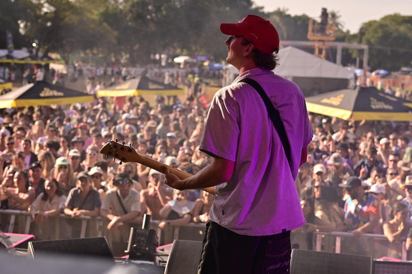 Guitarist in a red cap performs on stage at an outdoor concert, with a large, attentive crowd in the background under sunny skies.