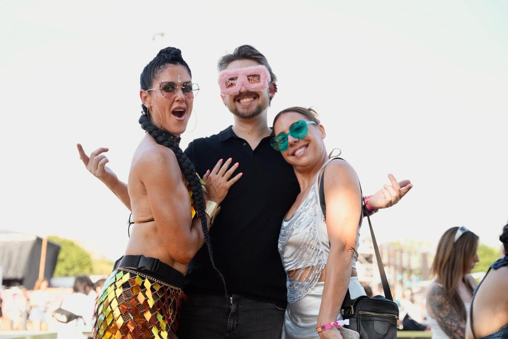 Three people posing outdoors, smiling and wearing colorful sunglasses. The person on the left has braided hair and a sequined outfit.