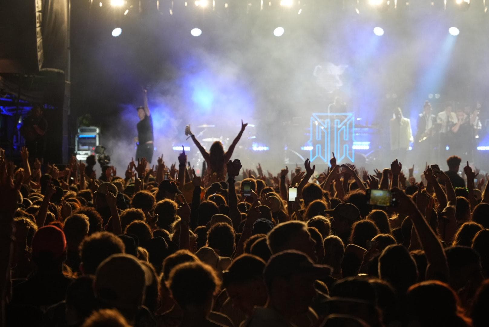 A lively concert crowd with raised hands, illuminated by stage lights and fog, with a performer on stage in the background.