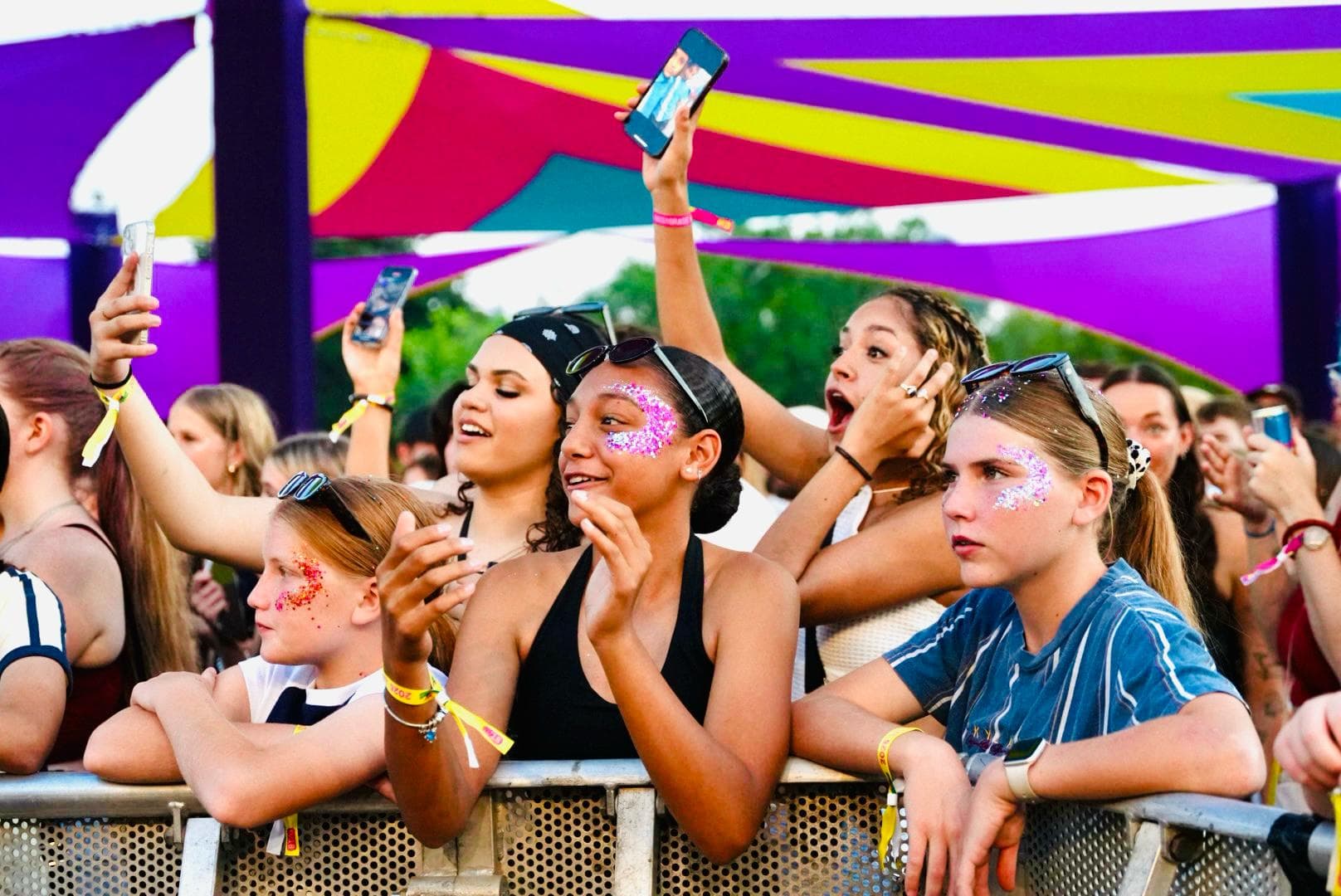 Audience of young people with glitter on faces, enjoying a concert, holding phones, and cheering under colorful canopy.