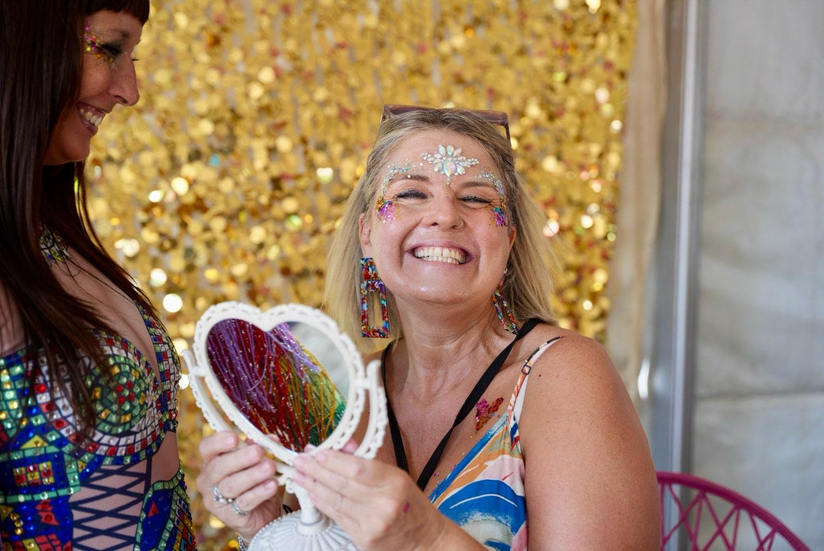 A woman with glittery face paint smiles while holding a heart-shaped mirror. Another person stands nearby. Background is a gold sequin curtain.