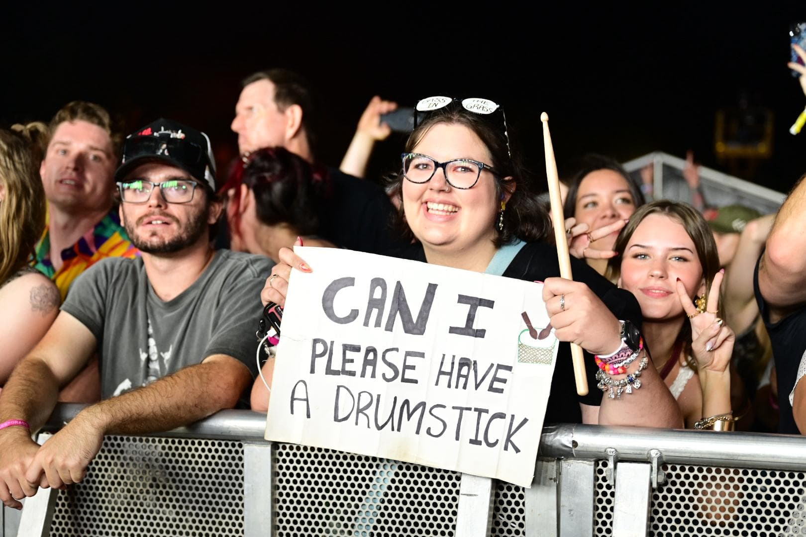 Concertgoers behind a barrier, one holding a sign that reads, "CAN I PLEASE HAVE A DRUMSTICK," smiling and gesturing with drumsticks.