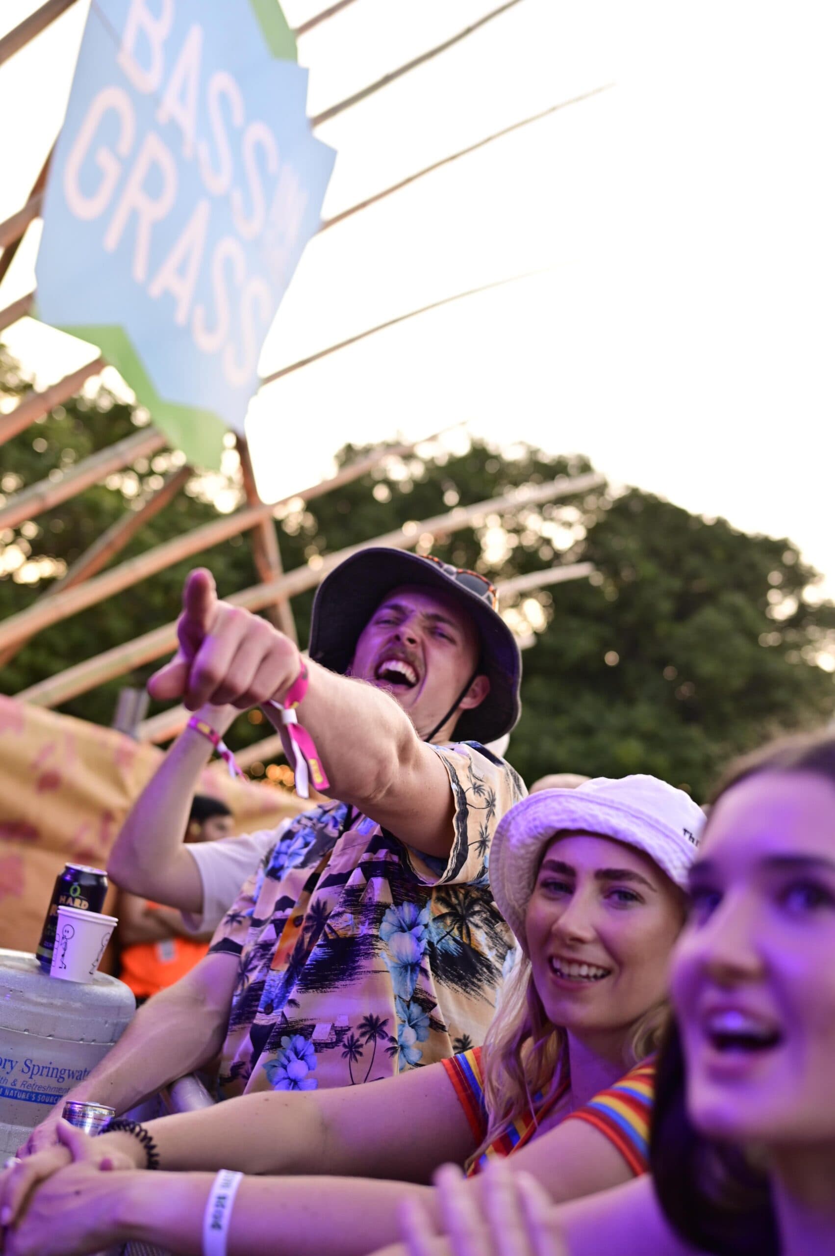 People enjoying a music festival, one pointing excitedly. They are wearing casual, colorful clothing and hats. A "Bass in Grass" sign is visible.