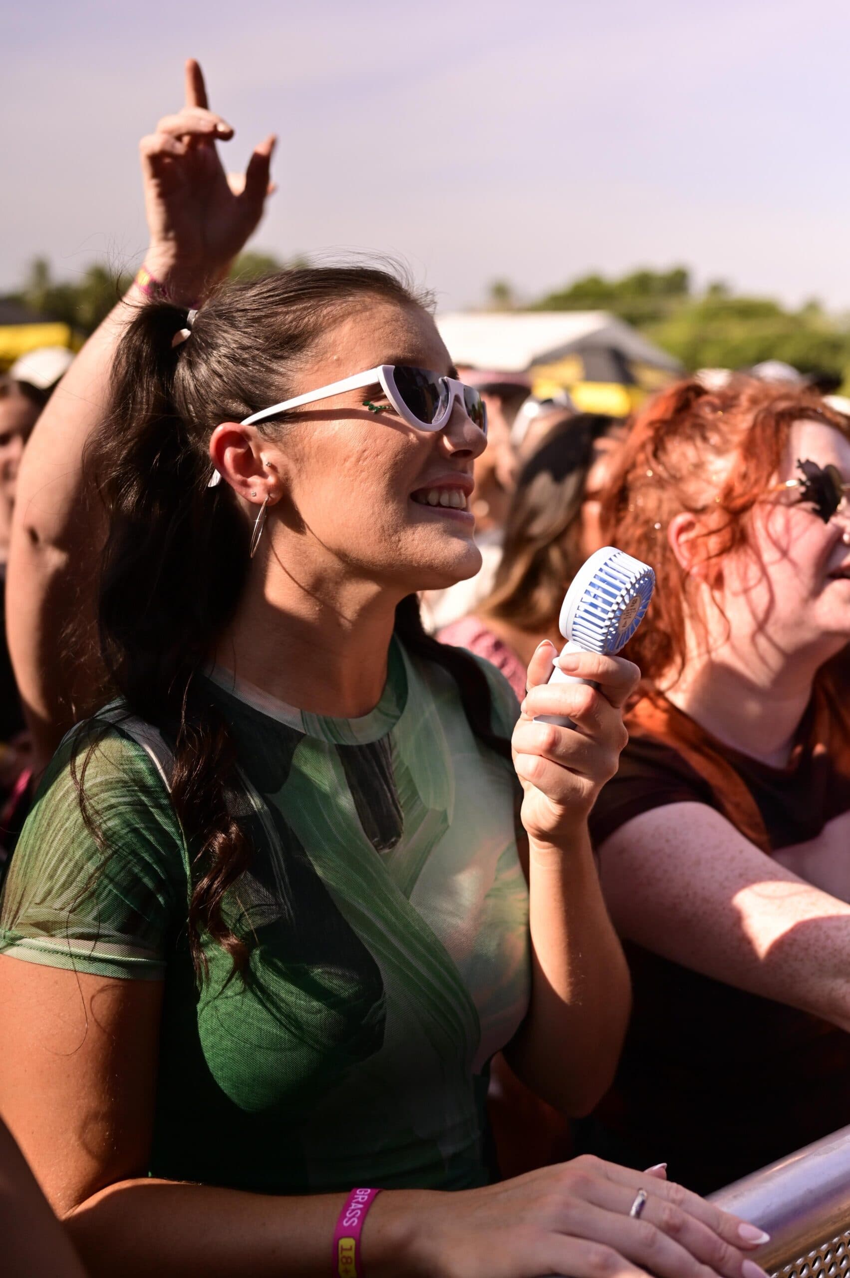 Woman in sunglasses smiles and holds a blue microphone at an outdoor event, surrounded by a crowd under a sunny sky.