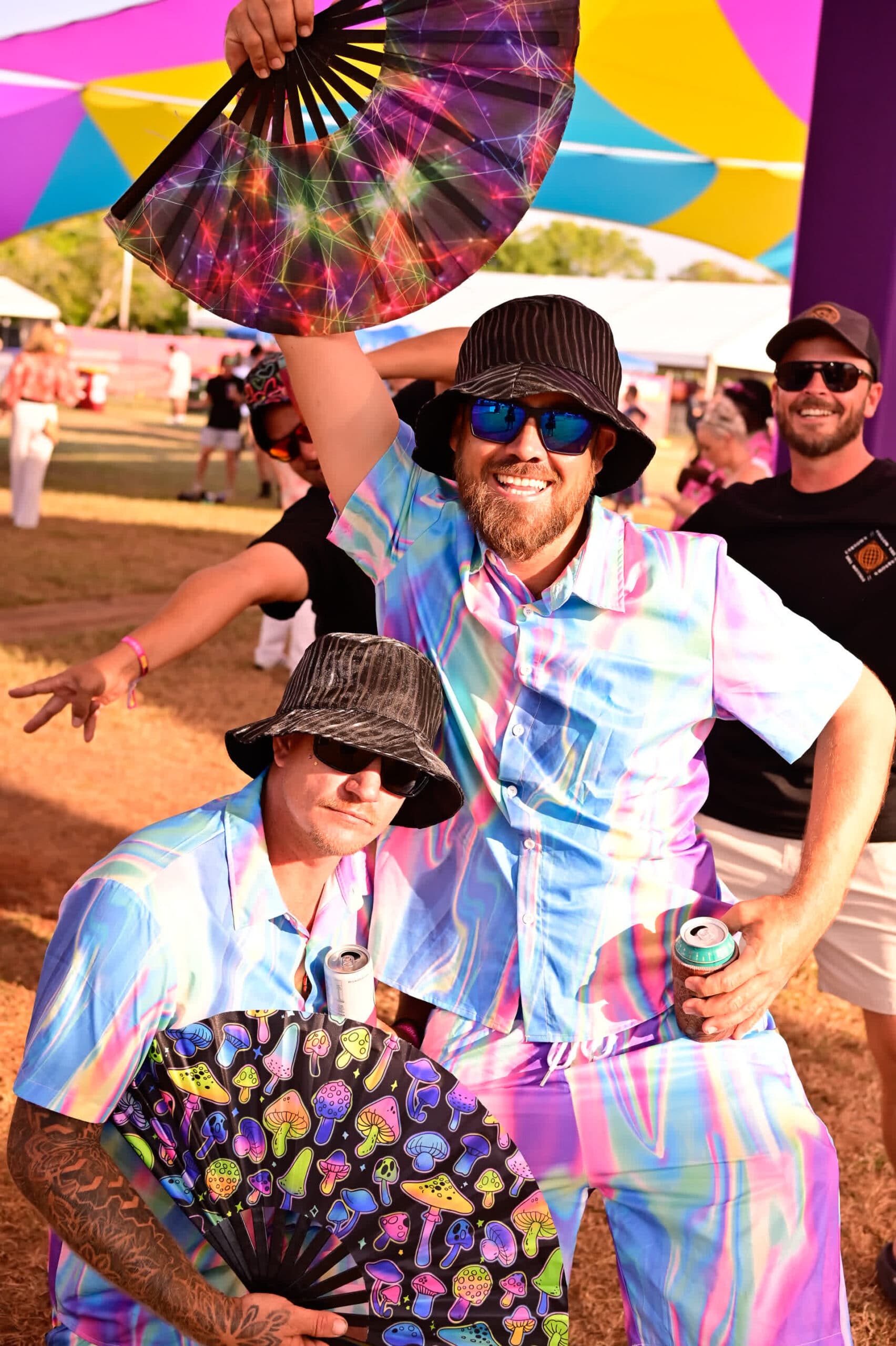 Two people in colorful outfits and bucket hats pose with fans at an outdoor event, with a vibrant canopy overhead and others in the background.