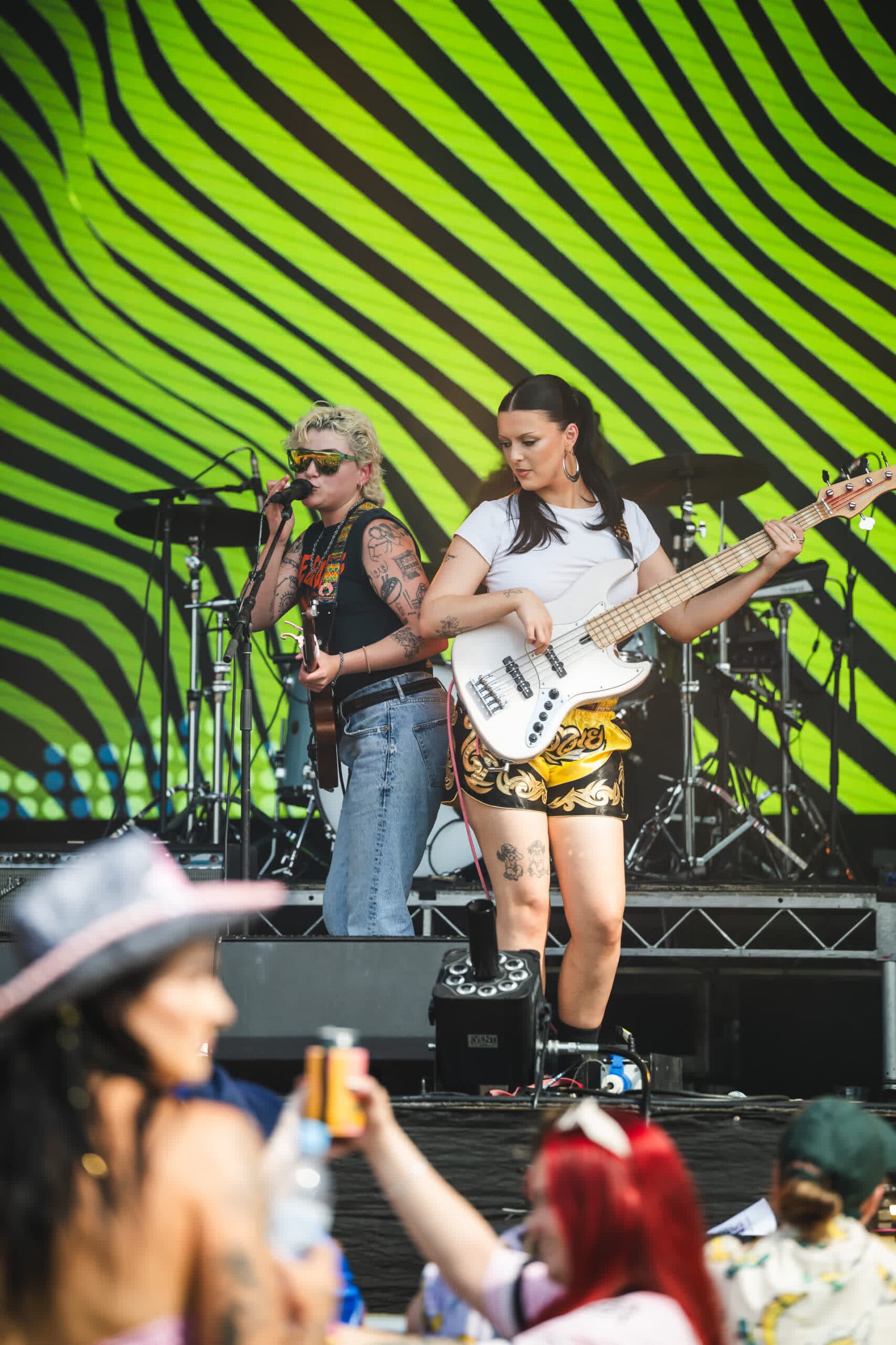 Two musicians perform on stage with a vibrant, wavy green and yellow backdrop. One plays bass guitar, the other sings into a microphone.