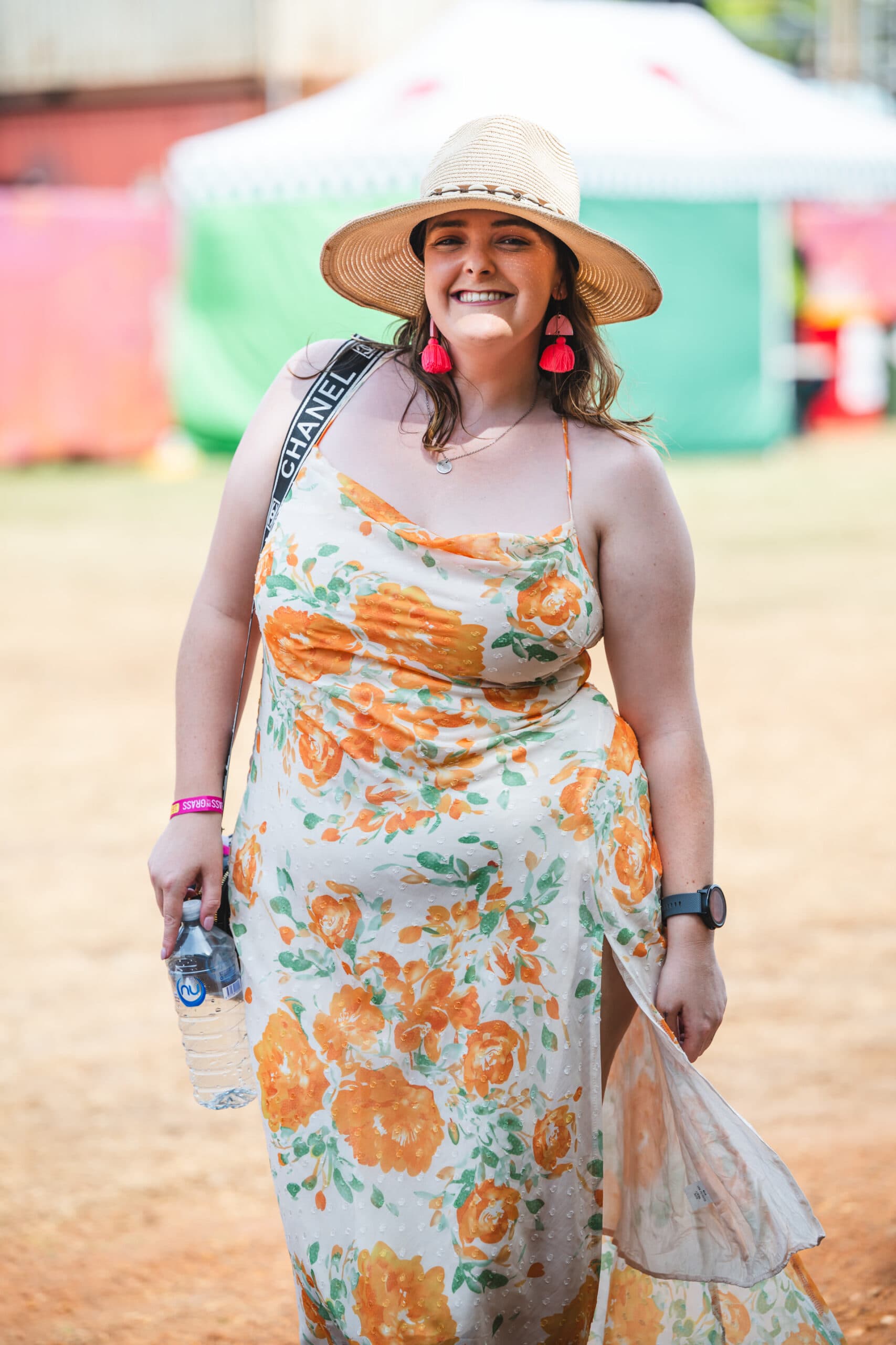 Woman in a floral dress and straw hat smiles, holding a water bottle. She wears pink earrings and a watch, standing outdoors on a sunny day.