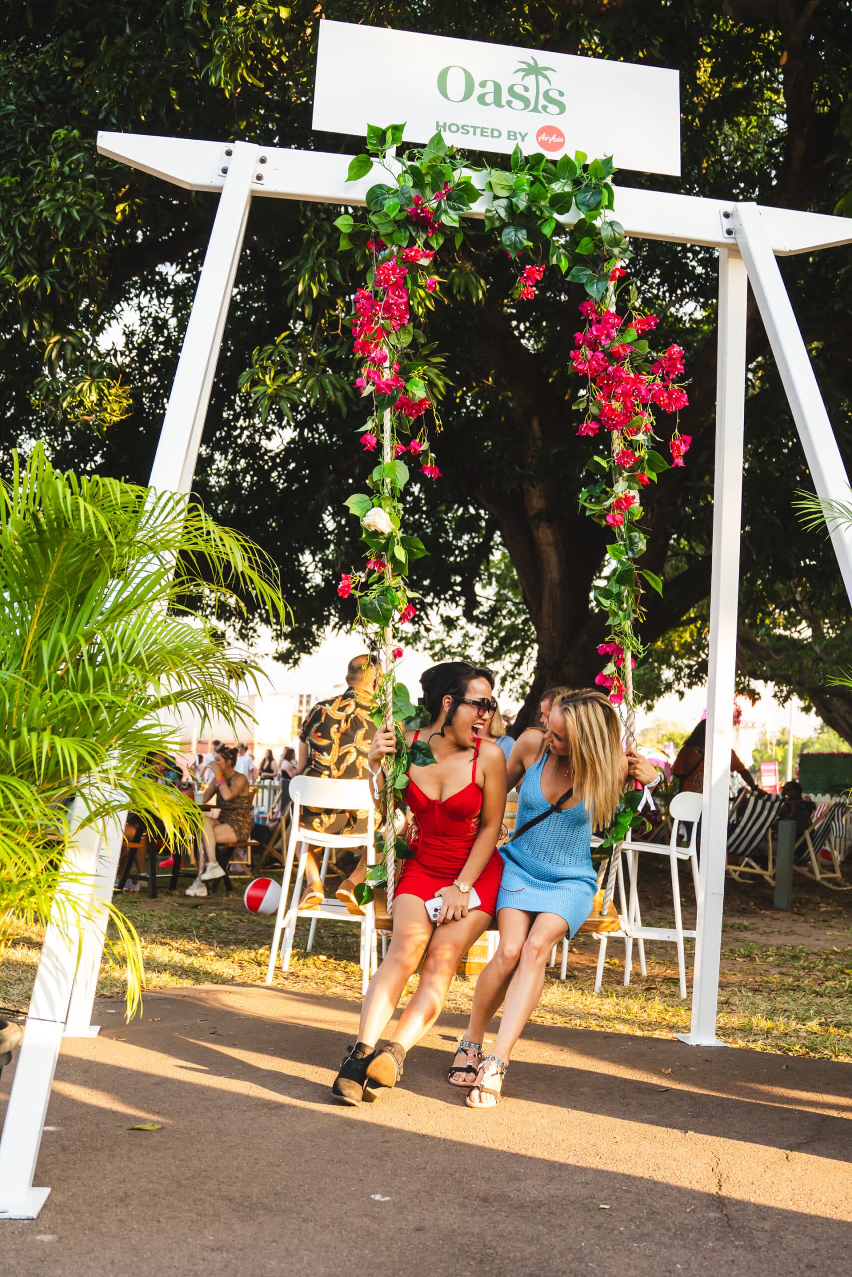 Two women sit on a floral-decorated swing under a tree at an outdoor event, surrounded by greenery and a crowd in the background.