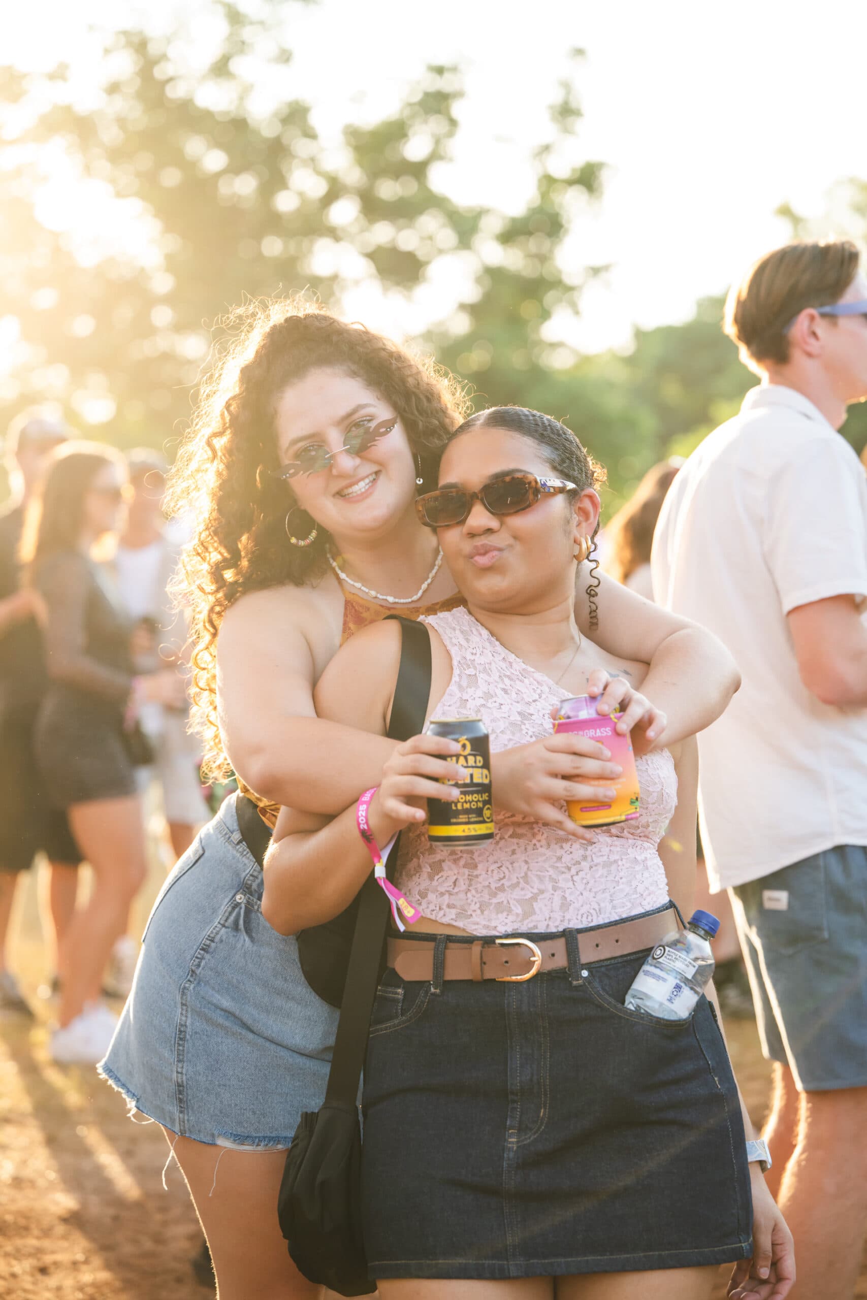 Two people enjoying an outdoor event, holding drinks, wearing sunglasses, with sunlight filtering through trees in the background.