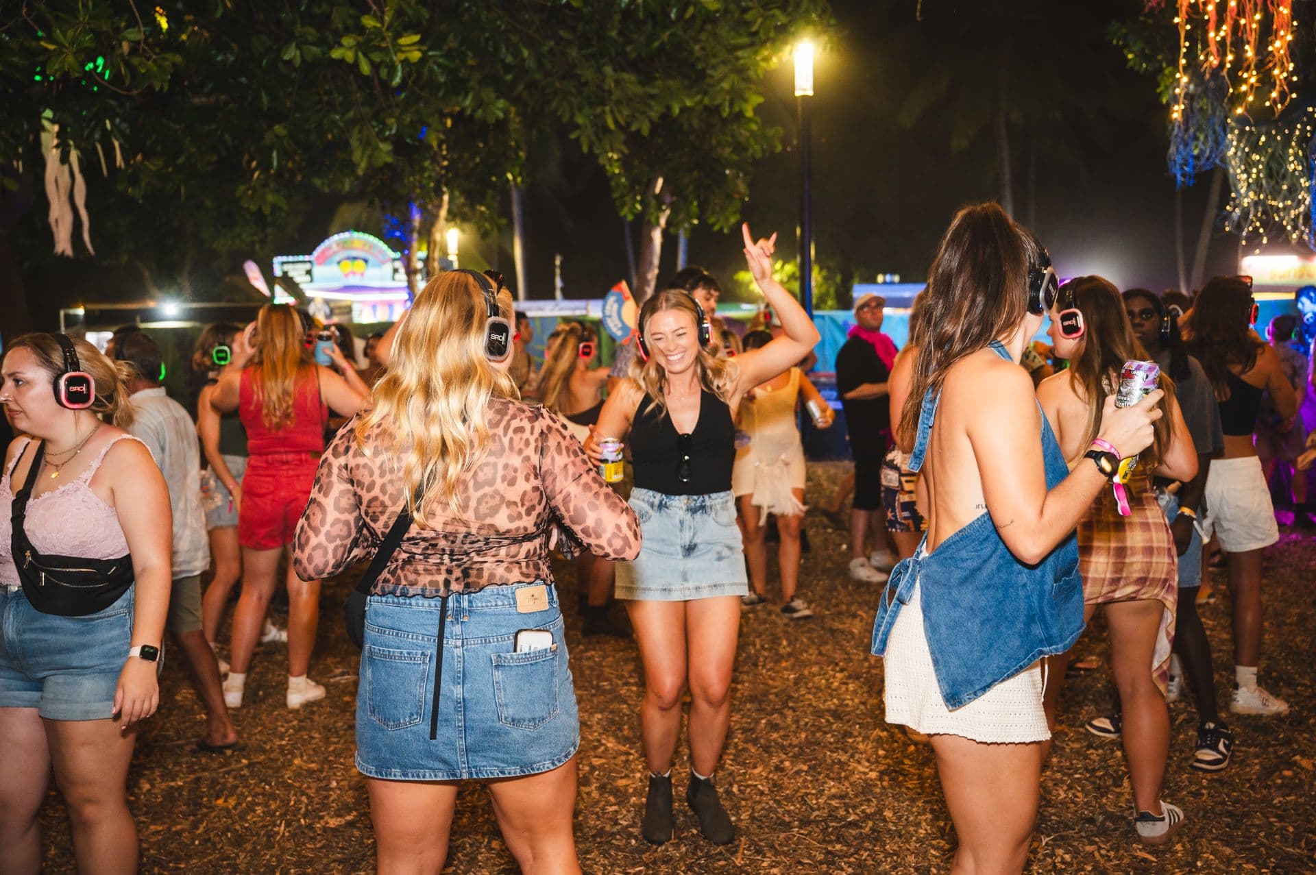 People dancing at an outdoor silent disco, wearing headphones, under colorful lights and trees. Casual summer attire and a lively atmosphere.