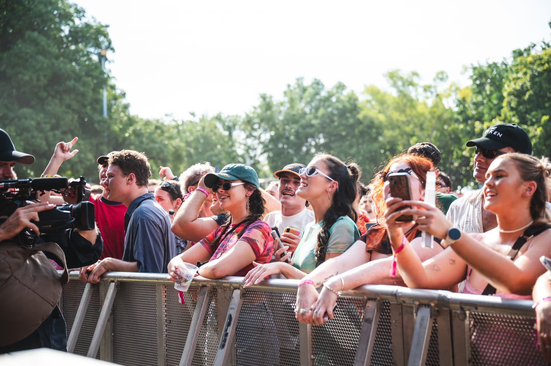 A lively crowd at an outdoor concert, smiling and cheering near the stage, with trees in the background on a sunny day.
