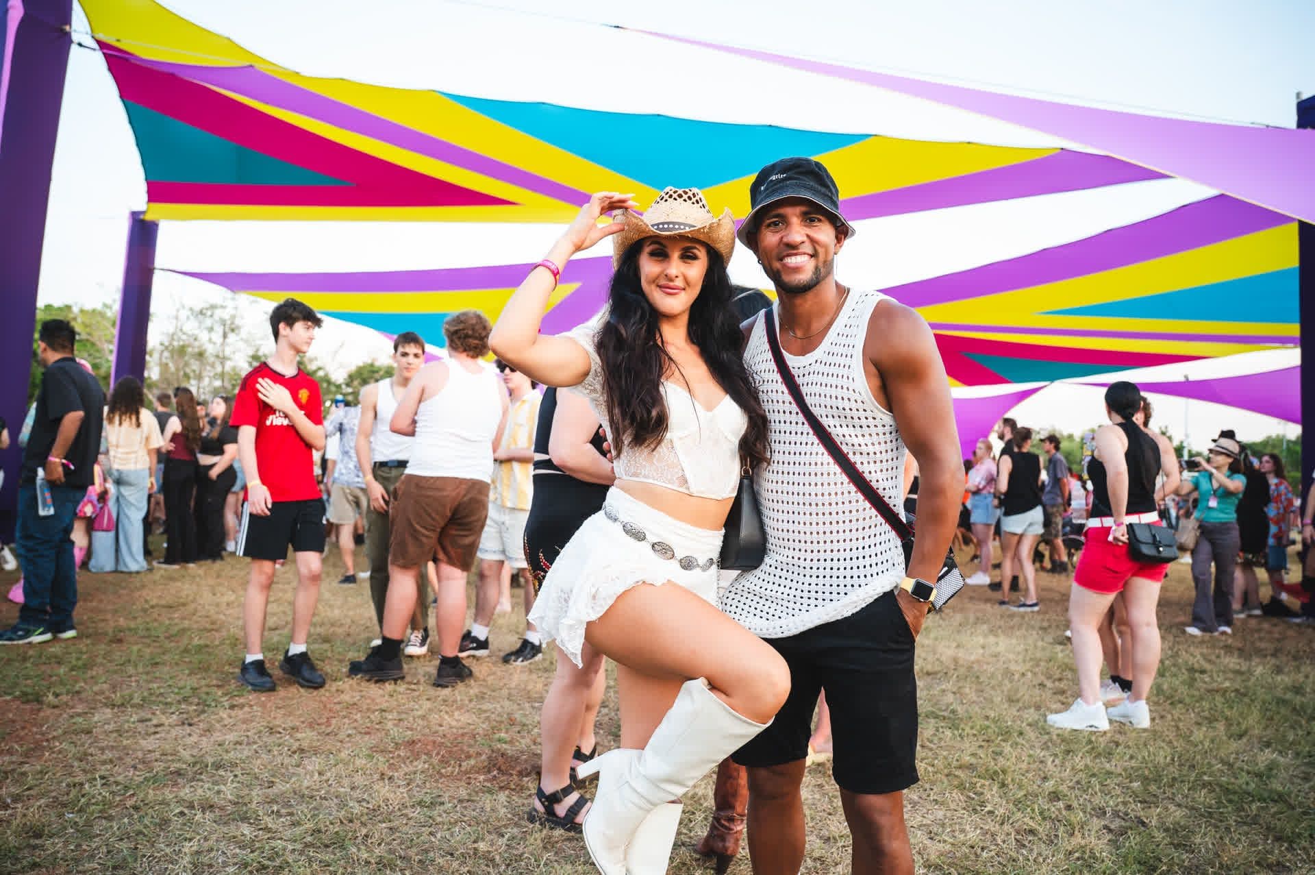 A smiling couple poses at an outdoor festival, surrounded by colorful canopy and people. The woman wears a cowboy hat and boots; the man wears a hat and tank top.