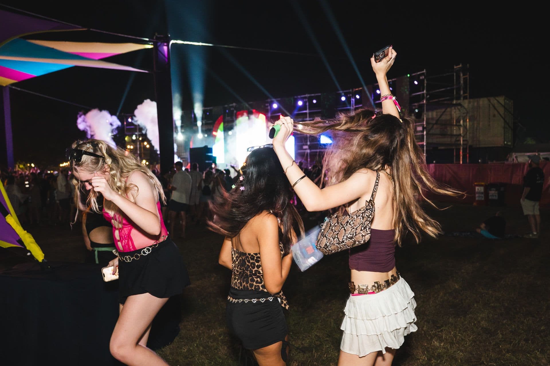 Three people dancing energetically at an outdoor music festival, with colourful lights and smoke in the background.