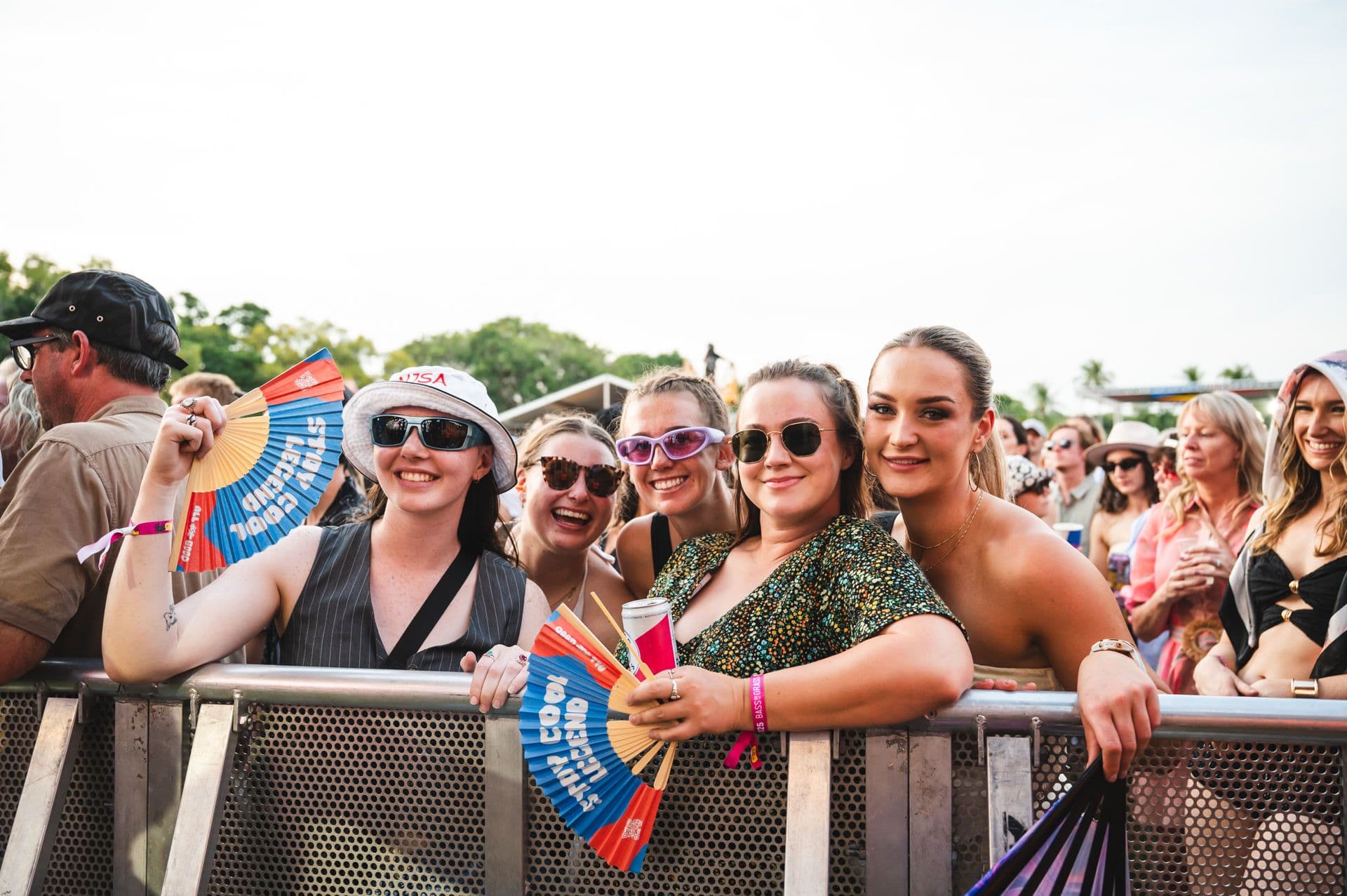 A group of people at an outdoor event, smiling and holding fans, stand behind a metal barrier. Trees and a crowd are visible in the background.