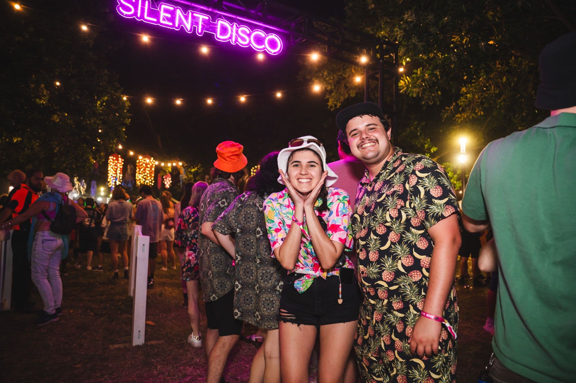 Two people smiling at a nighttime silent disco, wearing colorful shirts and hats, with a neon sign and festive lights in the background.
