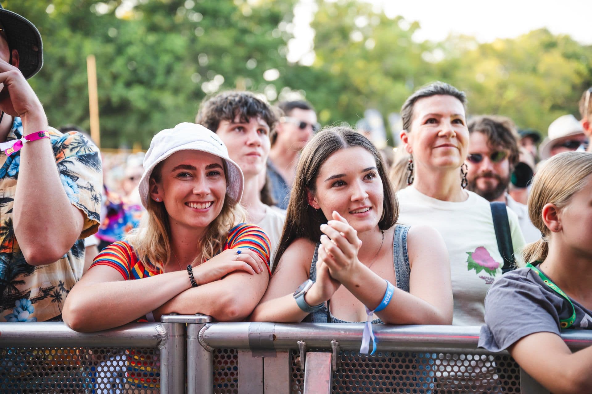 A crowd of people at an outdoor event, with two women in the foreground smiling and leaning on a metal barrier. Trees are visible in the background.