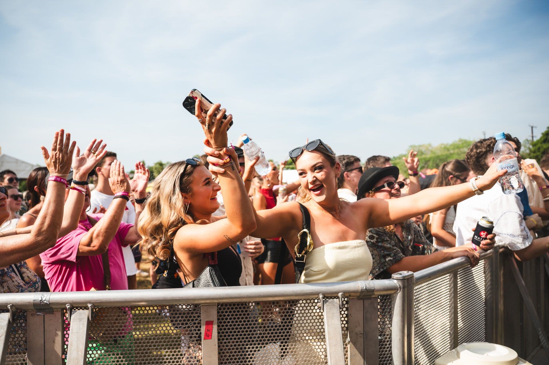 People enjoying a sunny outdoor concert, with two women in the foreground smiling and taking a selfie near the stage barrier.