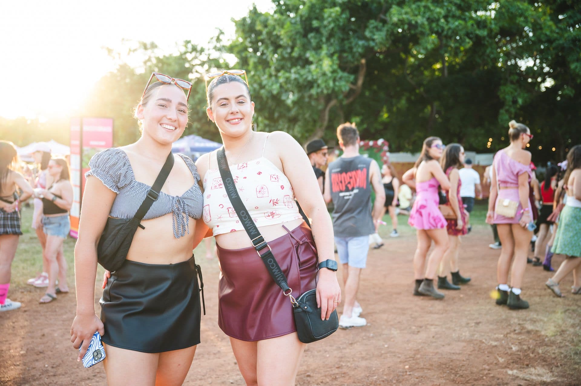 Two people smiling at an outdoor event, wearing casual summer outfits with crossbody bags. Other attendees and trees are visible in the background.