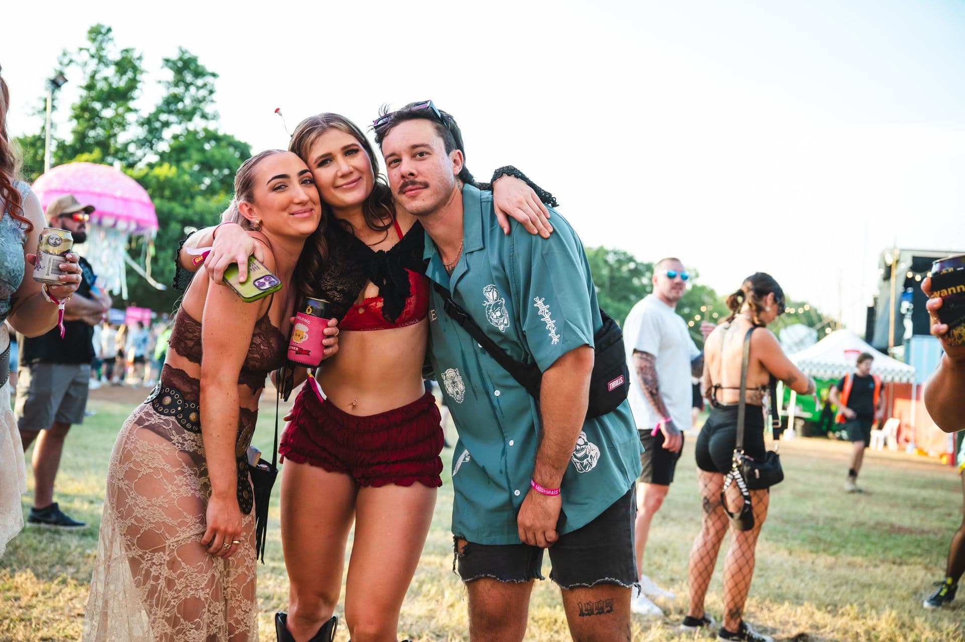 Three people posing and smiling at an outdoor festival, with colorful tents and other attendees in the background.