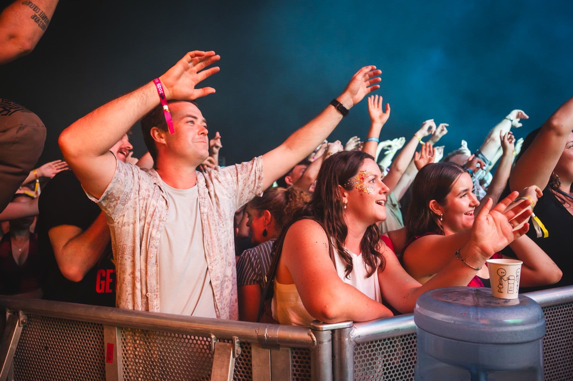 Enthusiastic crowd at a concert, people cheering and raising their hands near the stage barrier, with colorful lights in the background.