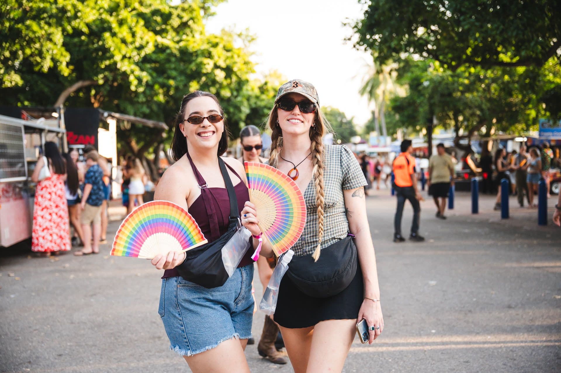 Two people smiling, holding rainbow fans at an outdoor event. They are wearing sunglasses and casual summer outfits, with trees and people in the background.
