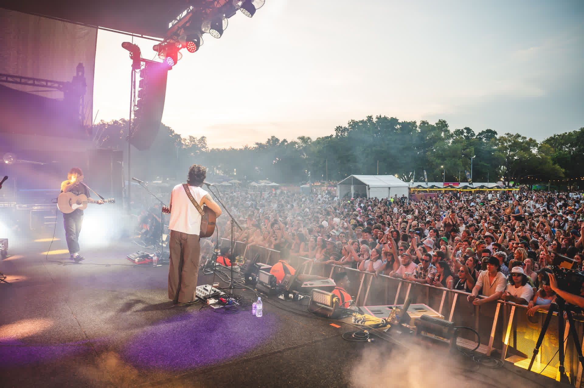 Musicians perform on an outdoor stage at sunset, facing a large, enthusiastic crowd. Stage lights and smoke create a vibrant atmosphere.