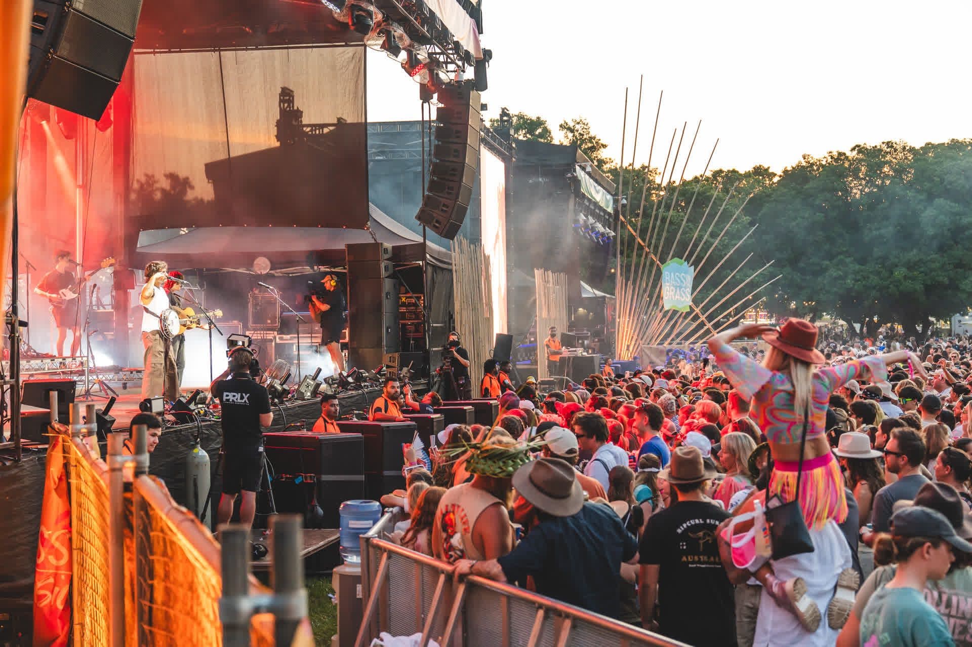 A vibrant outdoor music festival with a lively crowd, stage performers, colorful decorations, and a person on someone's shoulders enjoying the show.