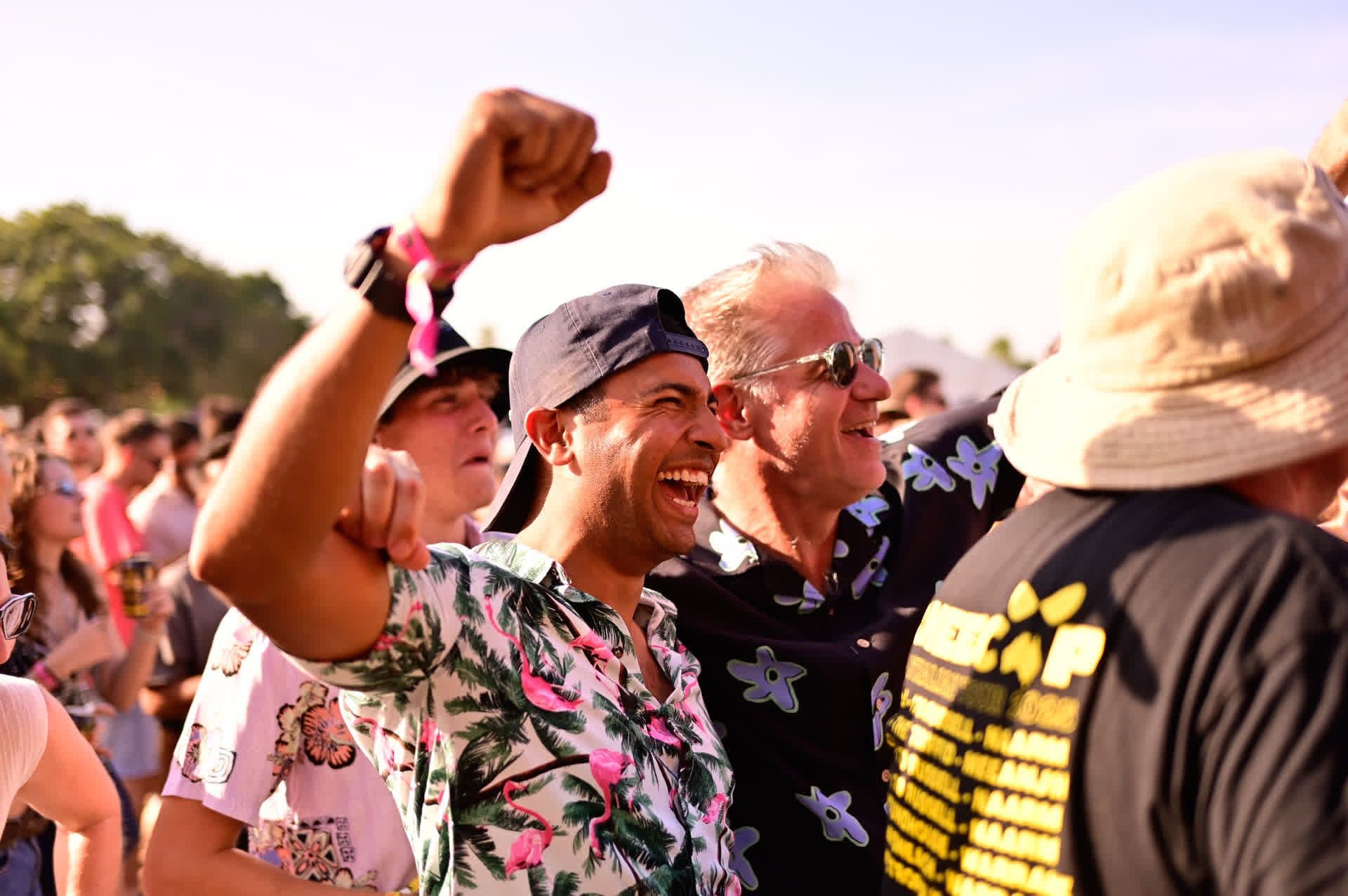 A crowd of people enjoying an outdoor event, with two men in floral shirts smiling and cheering. Sunny weather and clear skies in the background.