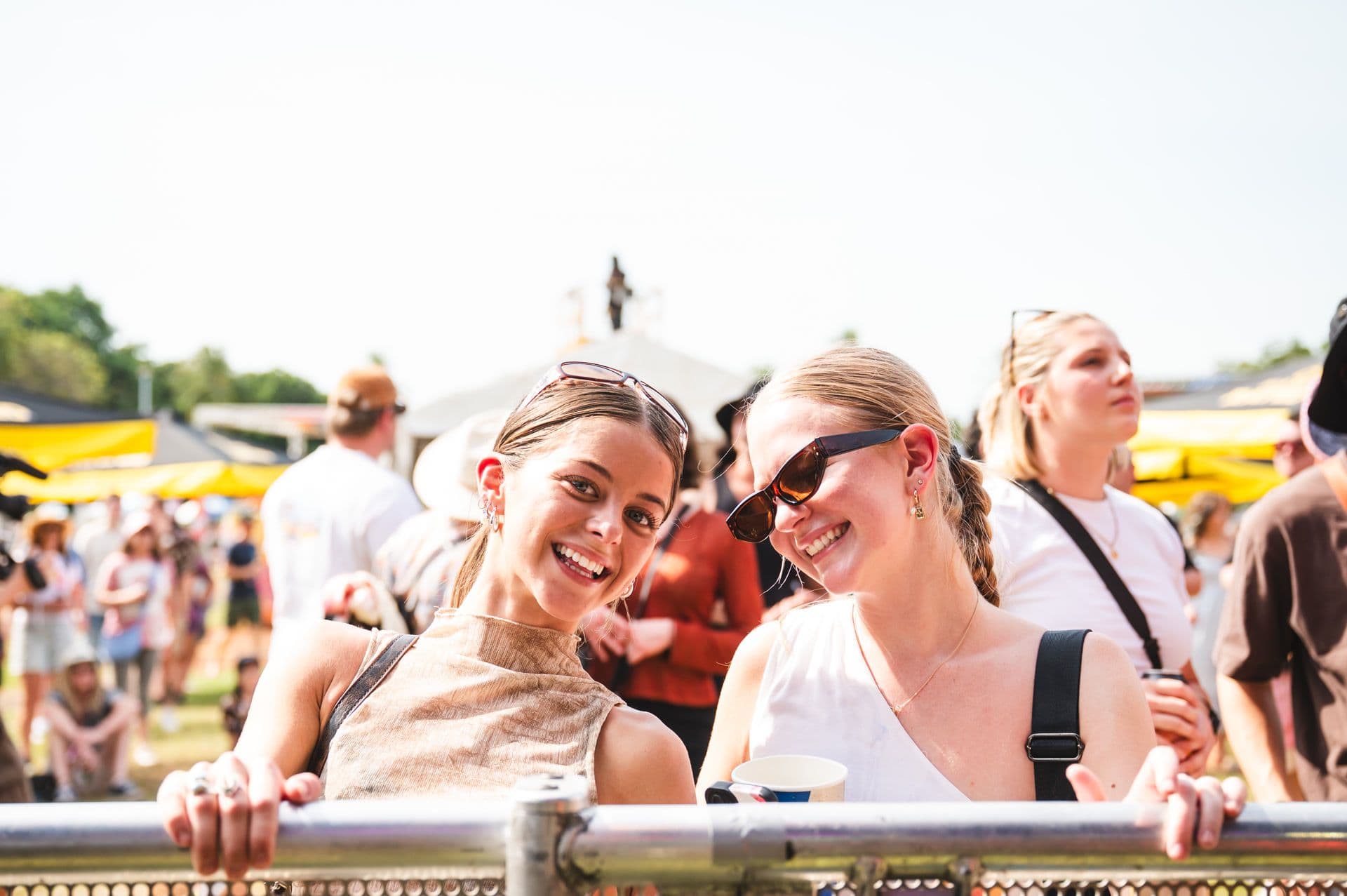 Two women smiling at an outdoor event, leaning on a metal barrier. One wears sunglasses; people and yellow umbrellas are in the background.